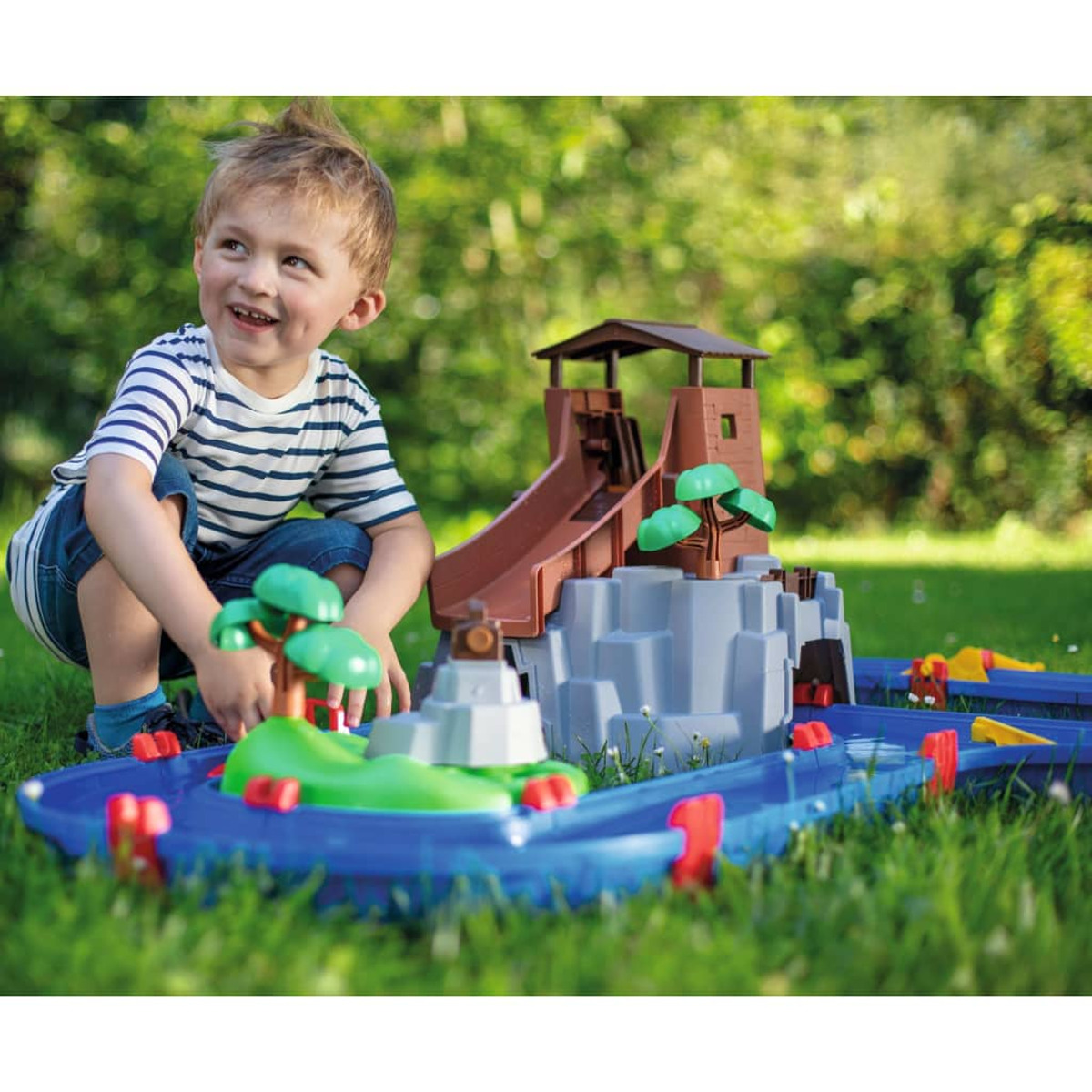 Ein kleiner Junge spielt mit einem blauen Spielzeug-Wasserpark auf grünem Gras.