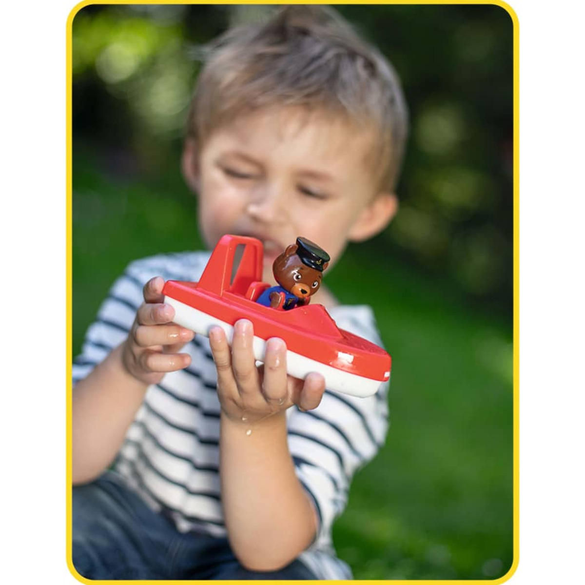 Ein Junge hält ein rot-weißes Spielzeugboot mit einer Bärenfigur. Der Hintergrund ist ein grüner, verschwommener Garten.