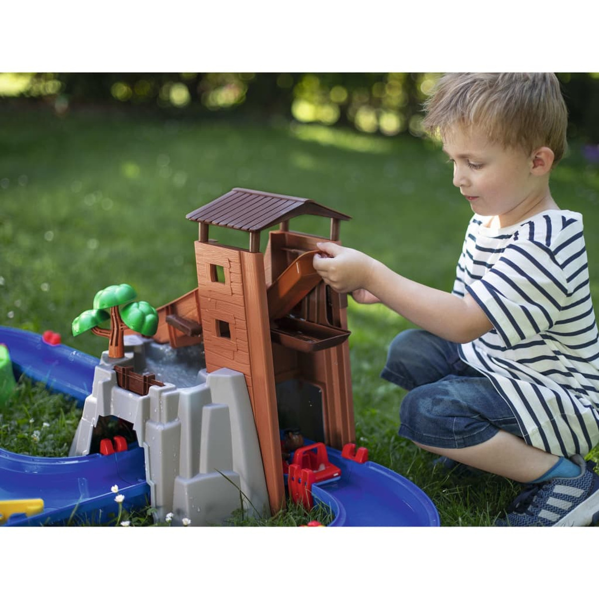 Ein Junge spielt mit einem Spielzeug-Wasserpark. Braune und graue Struktur, grüner Gras-Hintergrund.
