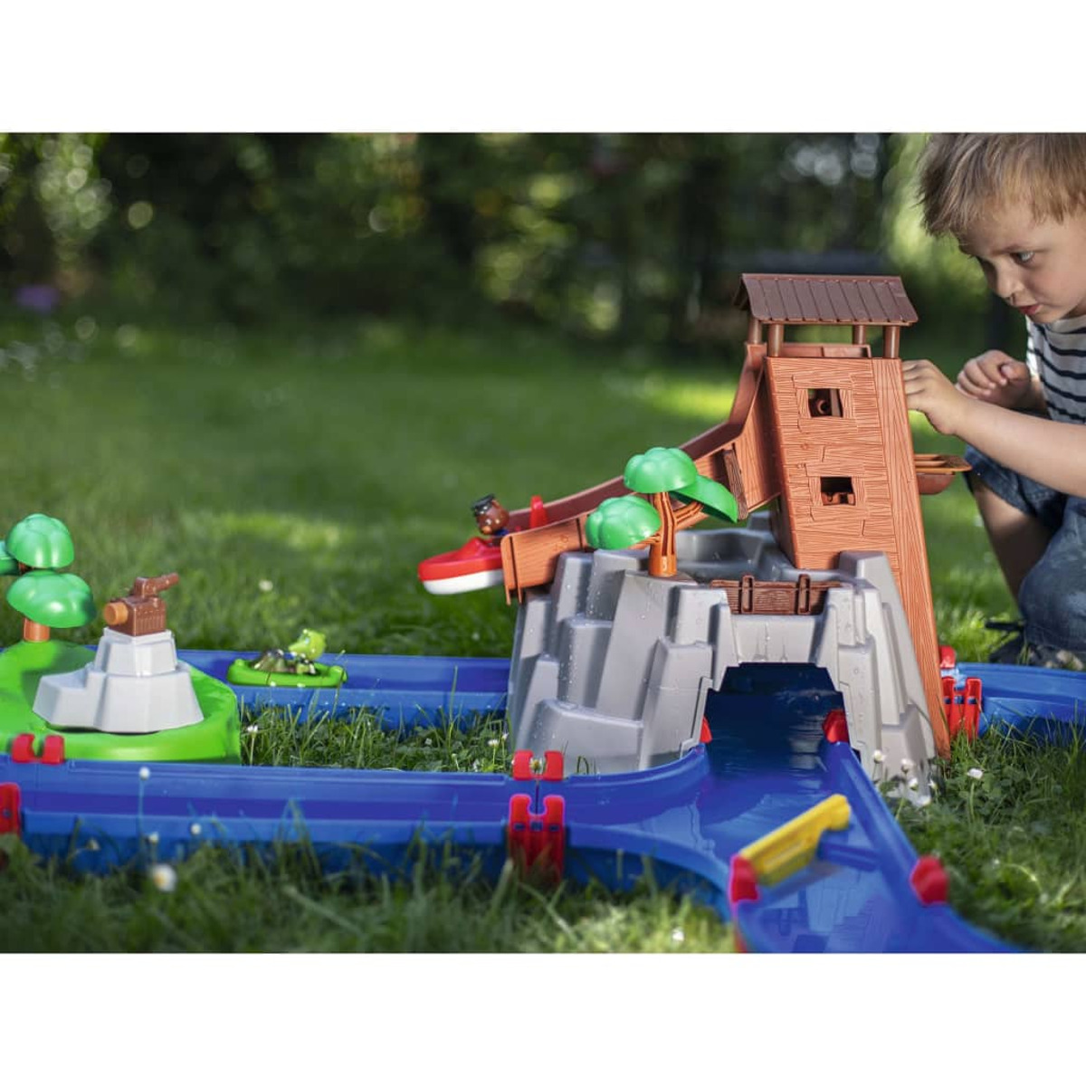Ein Kind spielt mit einem Spielzeug-Wasserpark mit Turm und Booten auf grünem Gras.