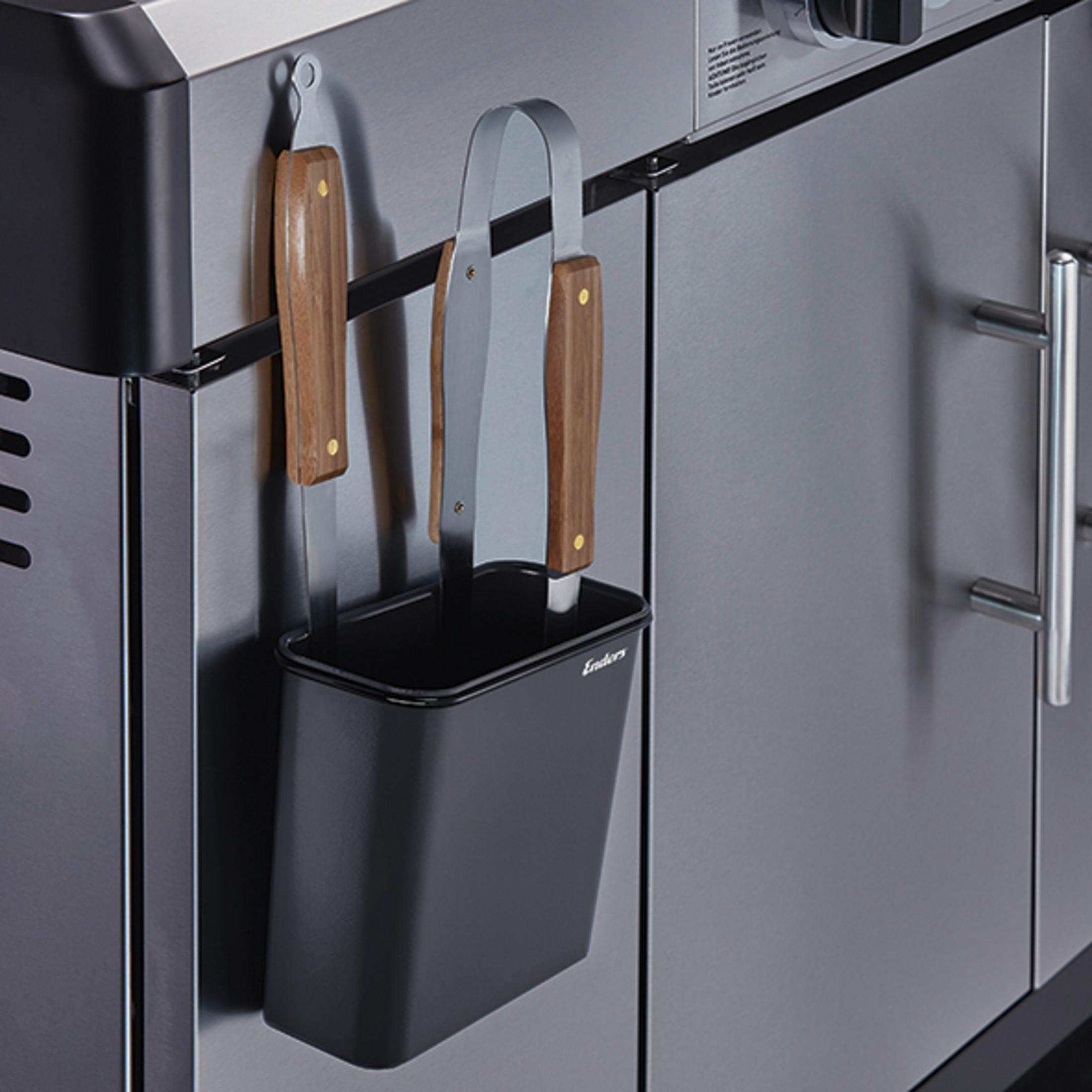 Edelstahlgrill mit hängenden Werkzeugen. Ein schwarzer Behälter enthält Zangen und Messer.