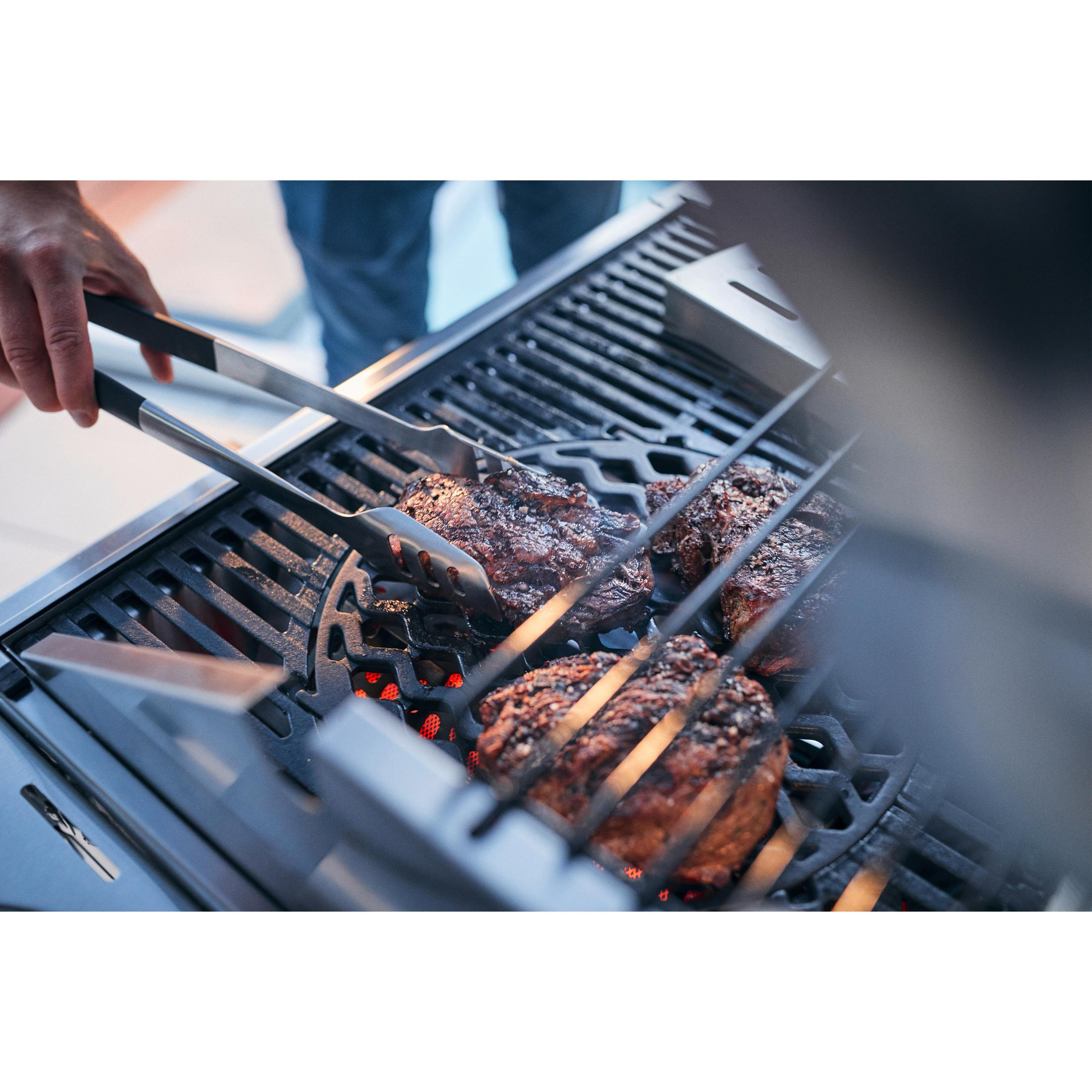 Nahaufnahme von grillenden Steaks auf einem Grill, Zange in der Hand, rot glühende Holzkohle.