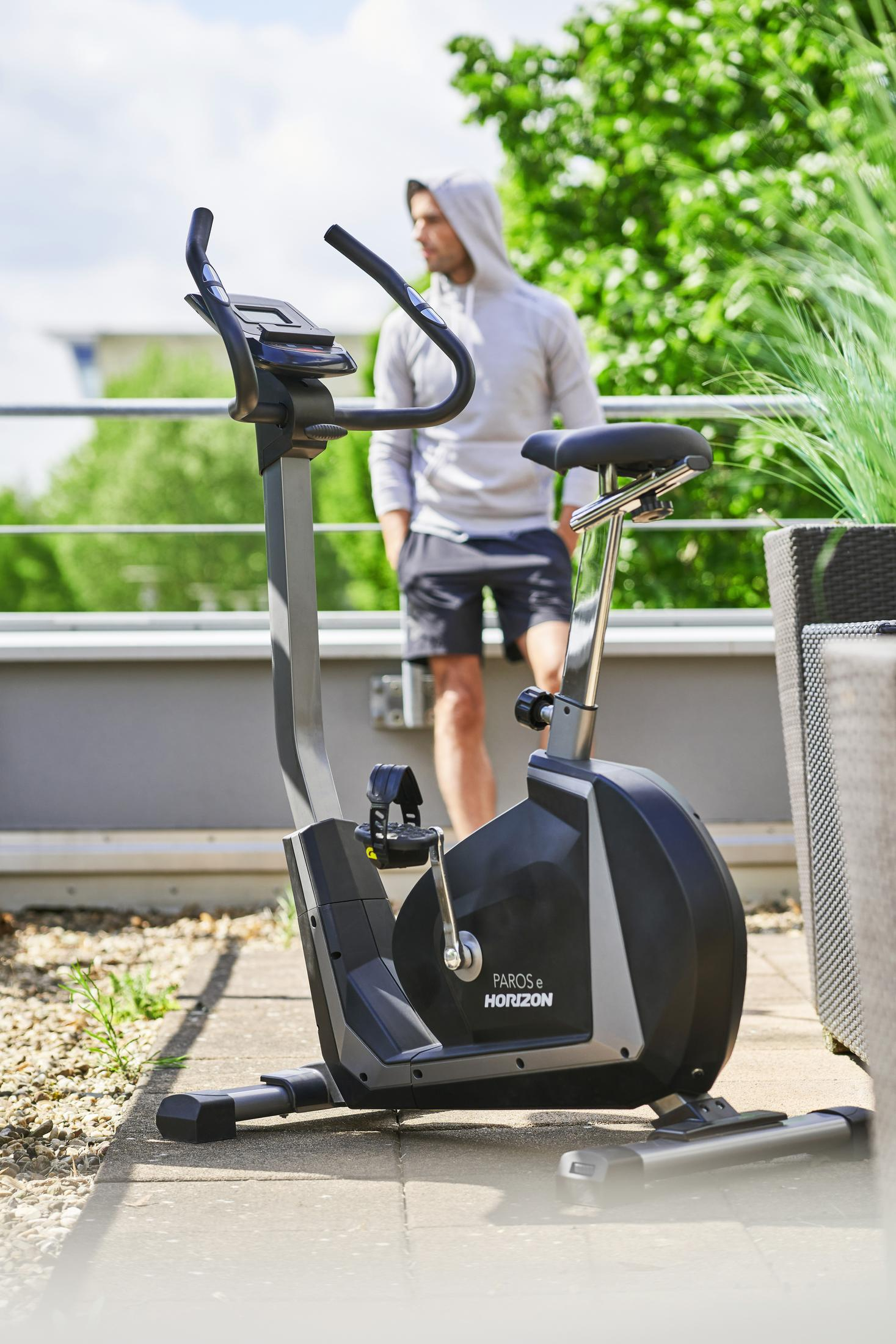 Een man met een hoodie staat bij een hometrainer op een dak. De fiets is zwart en grijs. Groene planten op de achtergrond.