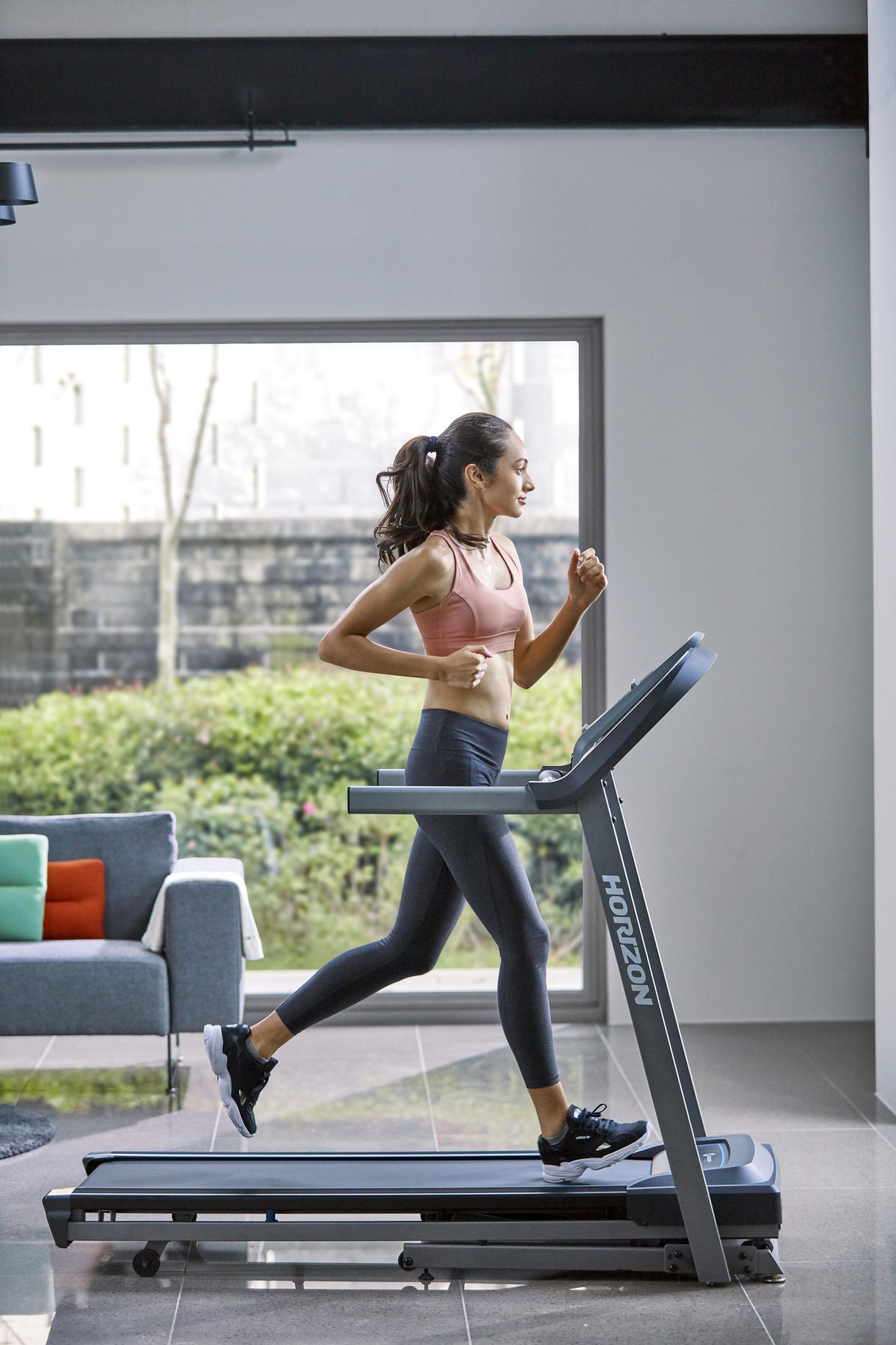 Kobieta biega na bieżni w domowej siłowni z oknem w tle.