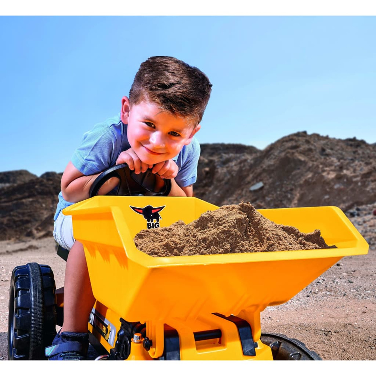 Chłopiec siedzi na żółtej zabawce ciężarówki wypełnionej piaskiem, uśmiechając się do kamery.