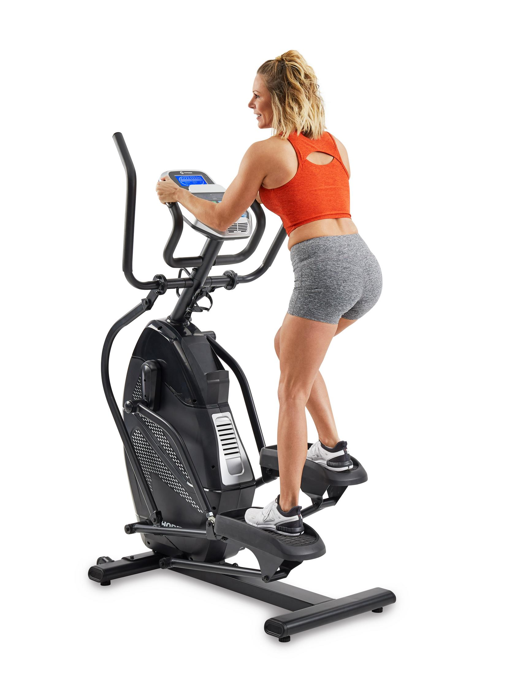 Mujer usando una máquina elíptica. Lleva un top naranja y pantalones cortos grises.