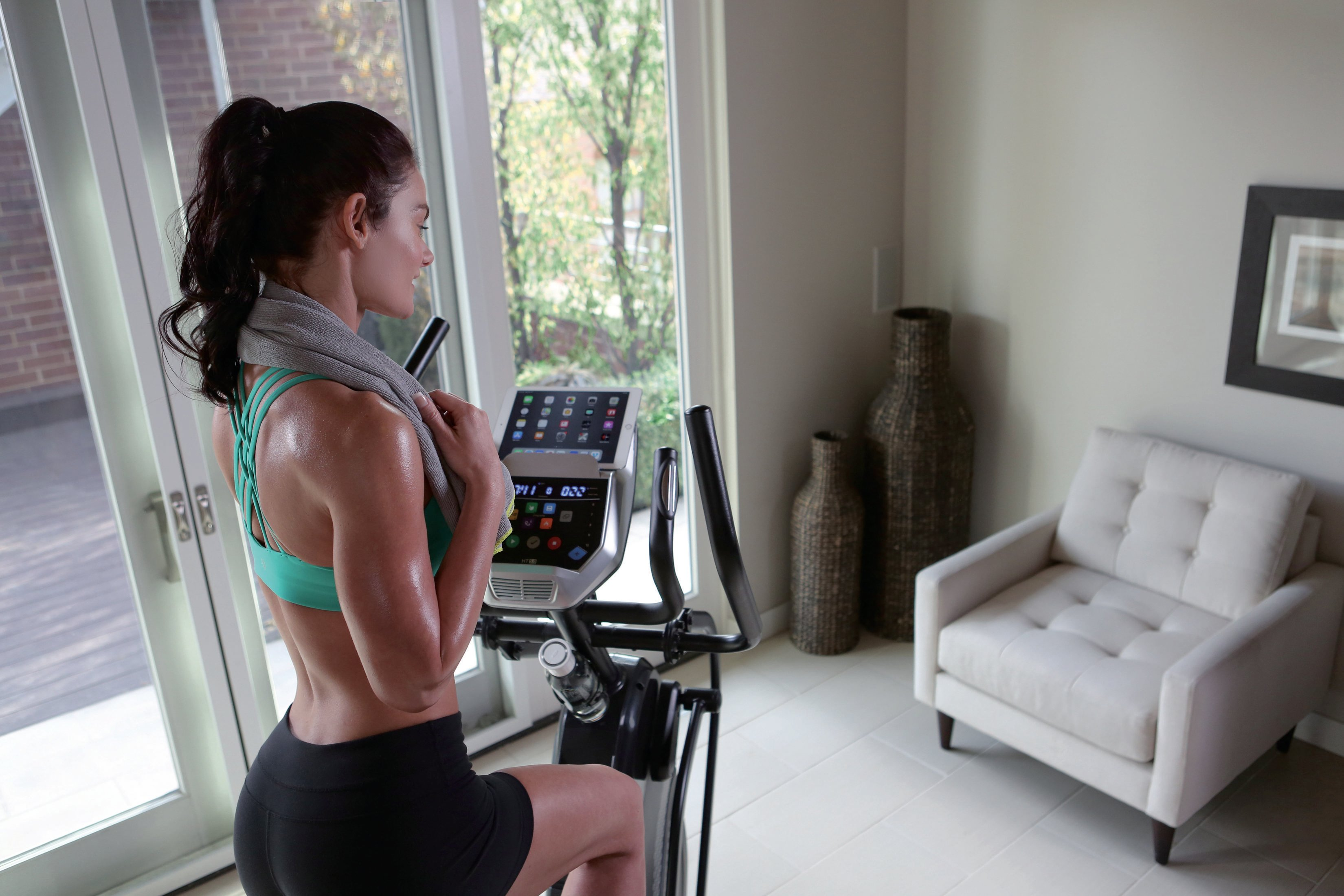 Mujer en una máquina elíptica, con una toalla alrededor del cuello, en un gimnasio en casa.