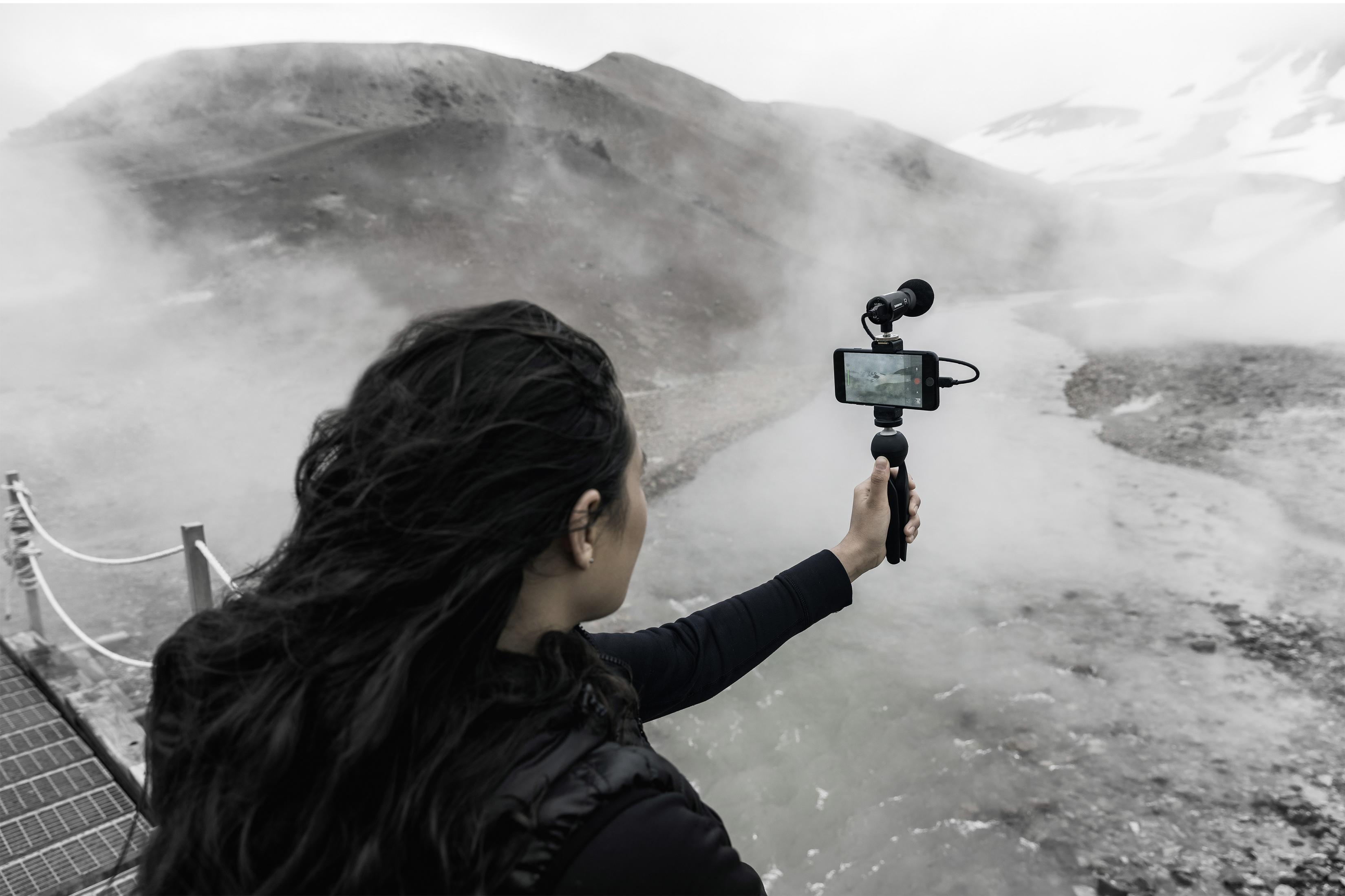 Frau filmt auf einer Brücke mit einem Handy und Mikrofon. Hintergrund ist eine neblige Landschaft.