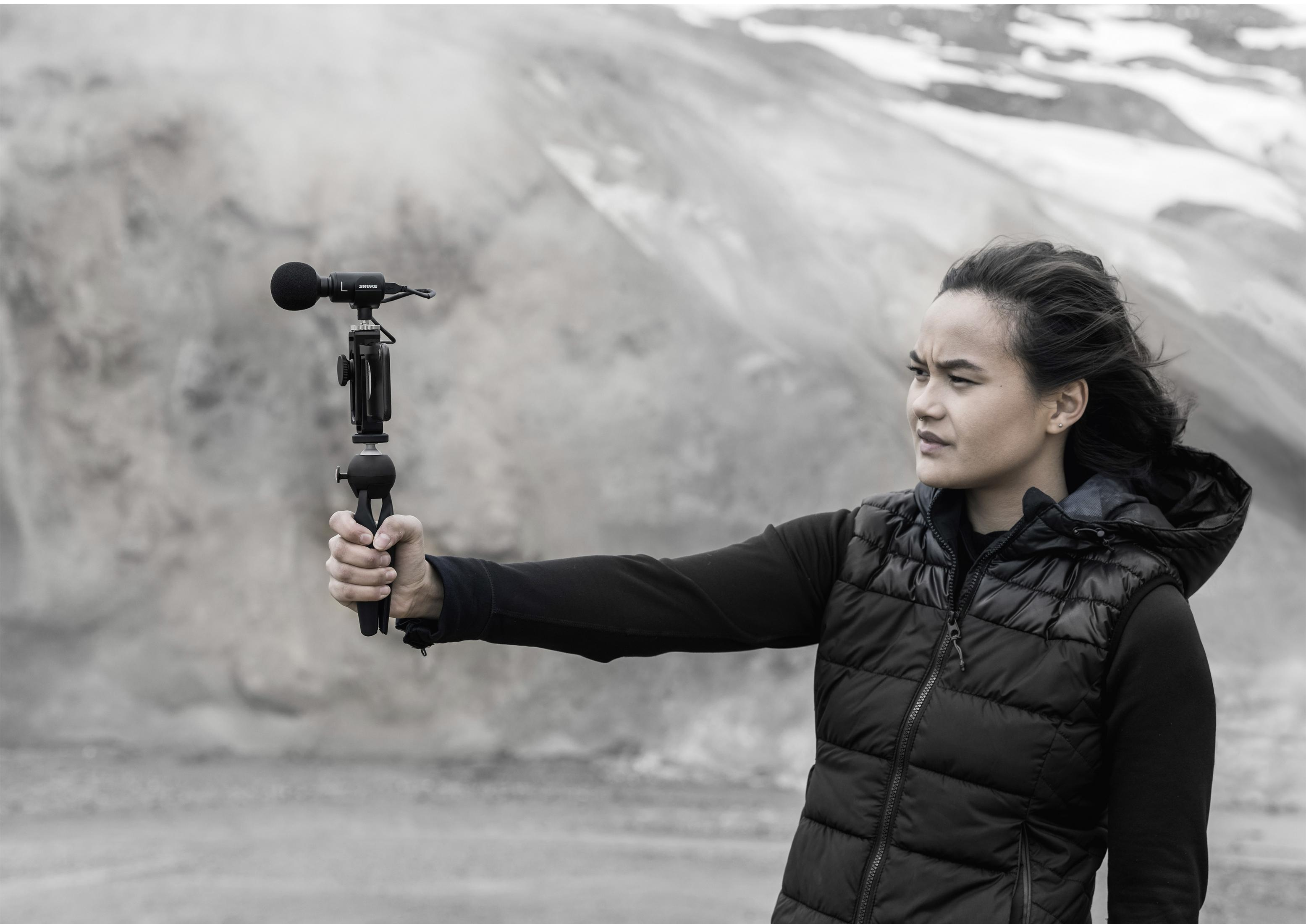 Frau hält Mikrofon, filmt mit Kamera, in bergiger Landschaft.