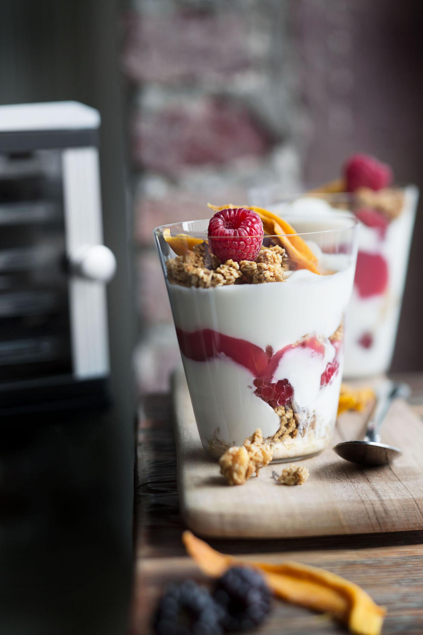 Due bicchieri di parfait allo yogurt con granola, frutti di bosco e mango. Sono posizionati su una tavola di legno.