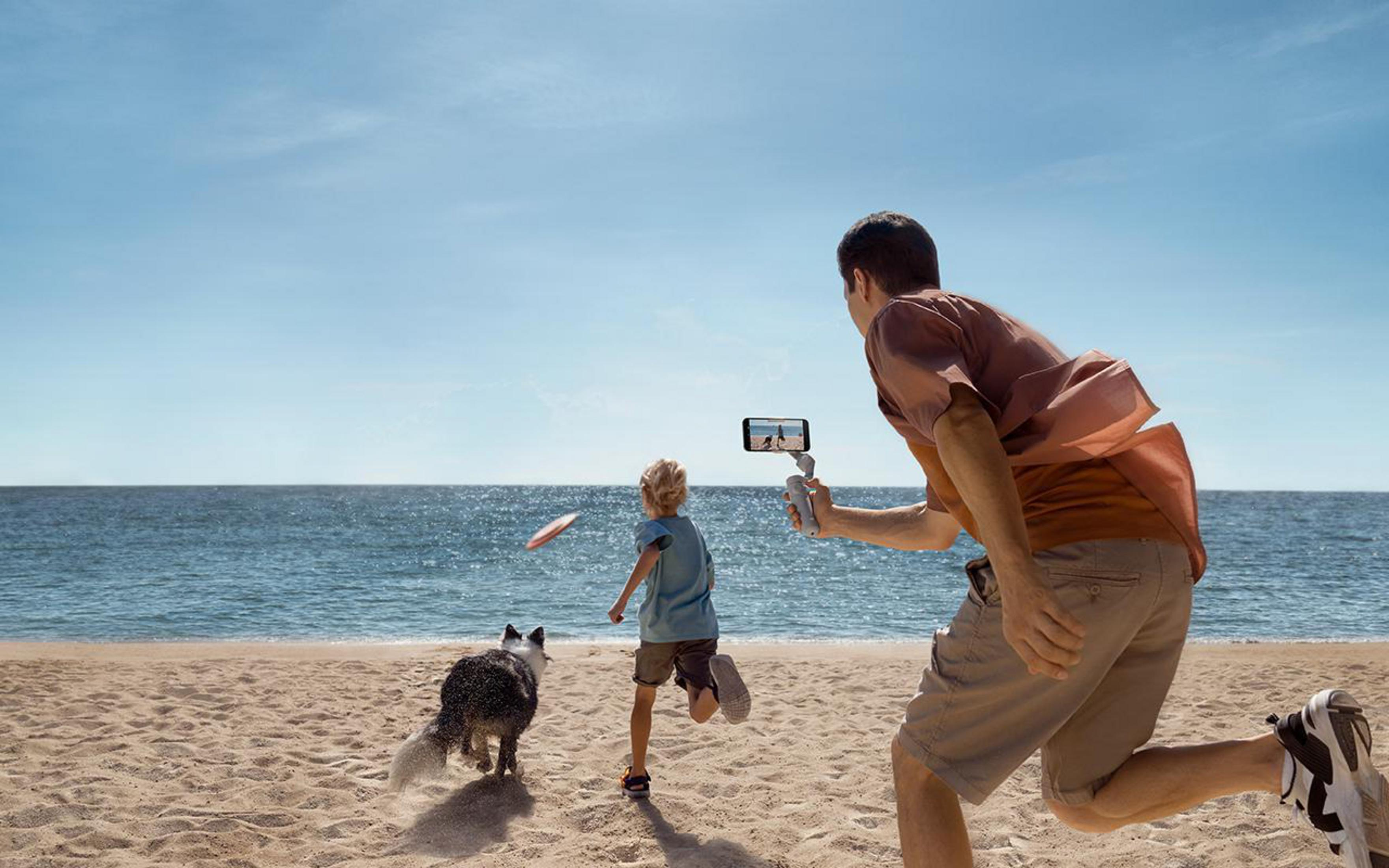Mann mit Telefon, der ein Video aufnimmt. Junge rennt mit Hund am Strand. Blaues Meer und Himmel.