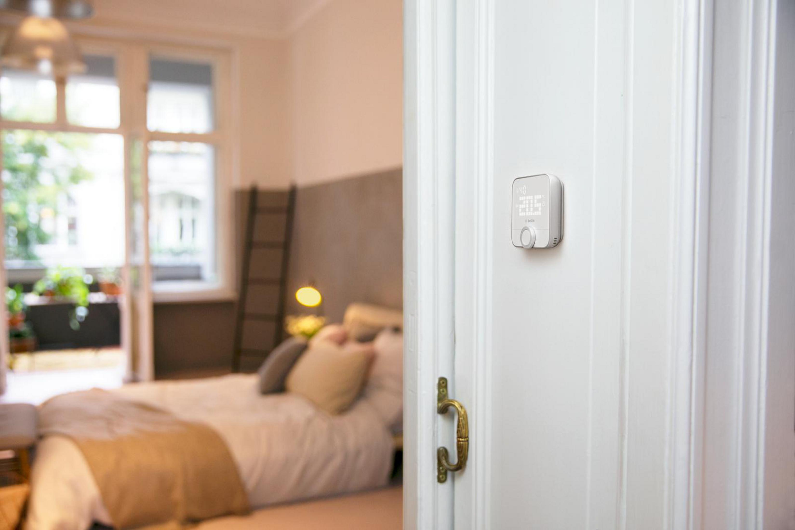 Eine weiße Wand mit einem Thermostat und einer Tür mit Griff, mit einem verschwommenen Schlafzimmer im Hintergrund.