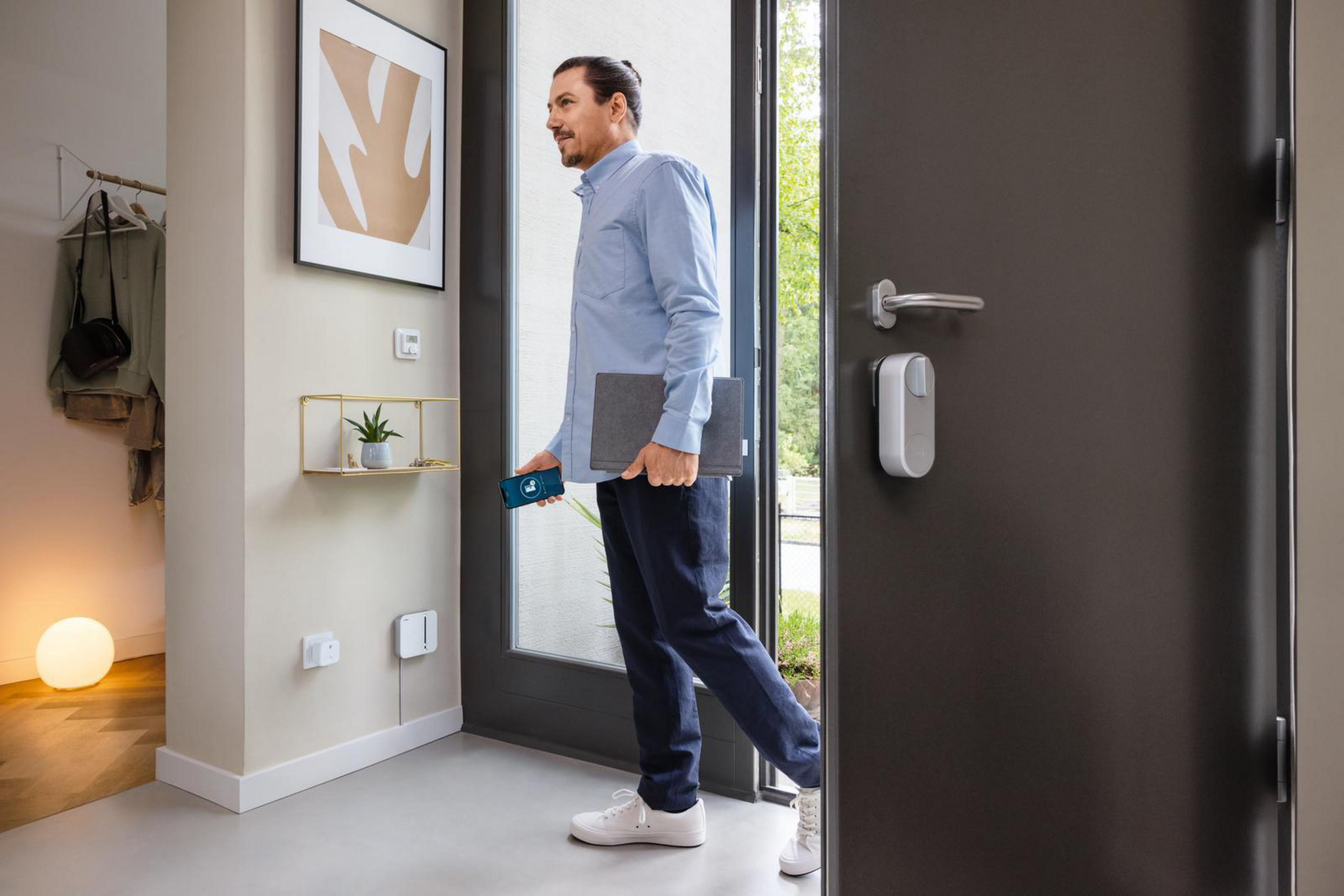 Ein Mann verlässt das Haus mit Laptop und Handy. Smart Lock an der Tür. Kleines Regal an der Wand.