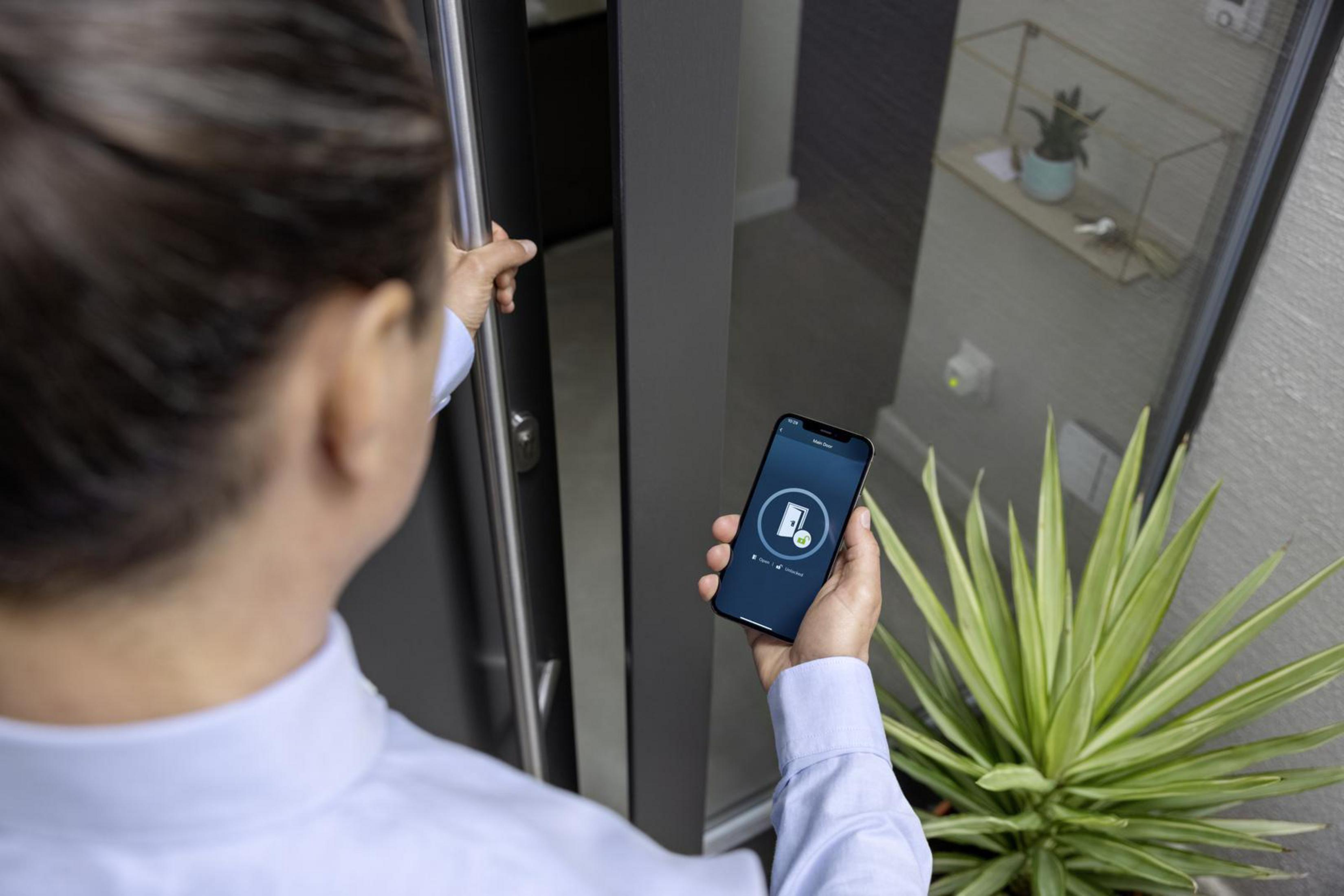 Eine Person entriegelt eine Tür mit einem Telefon. Eine Zimmerpflanze und ein Fenster sind sichtbar.