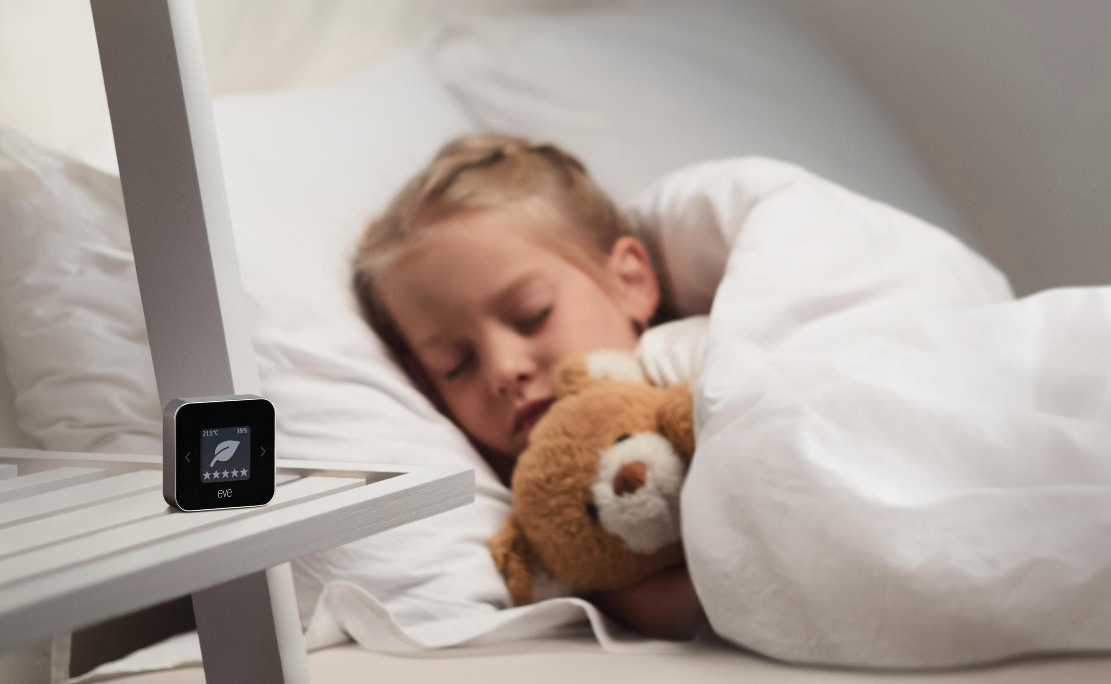 Dziecko śpi w łóżku, przytulając misia. Urządzenie z ekranem na stoliku nocnym.