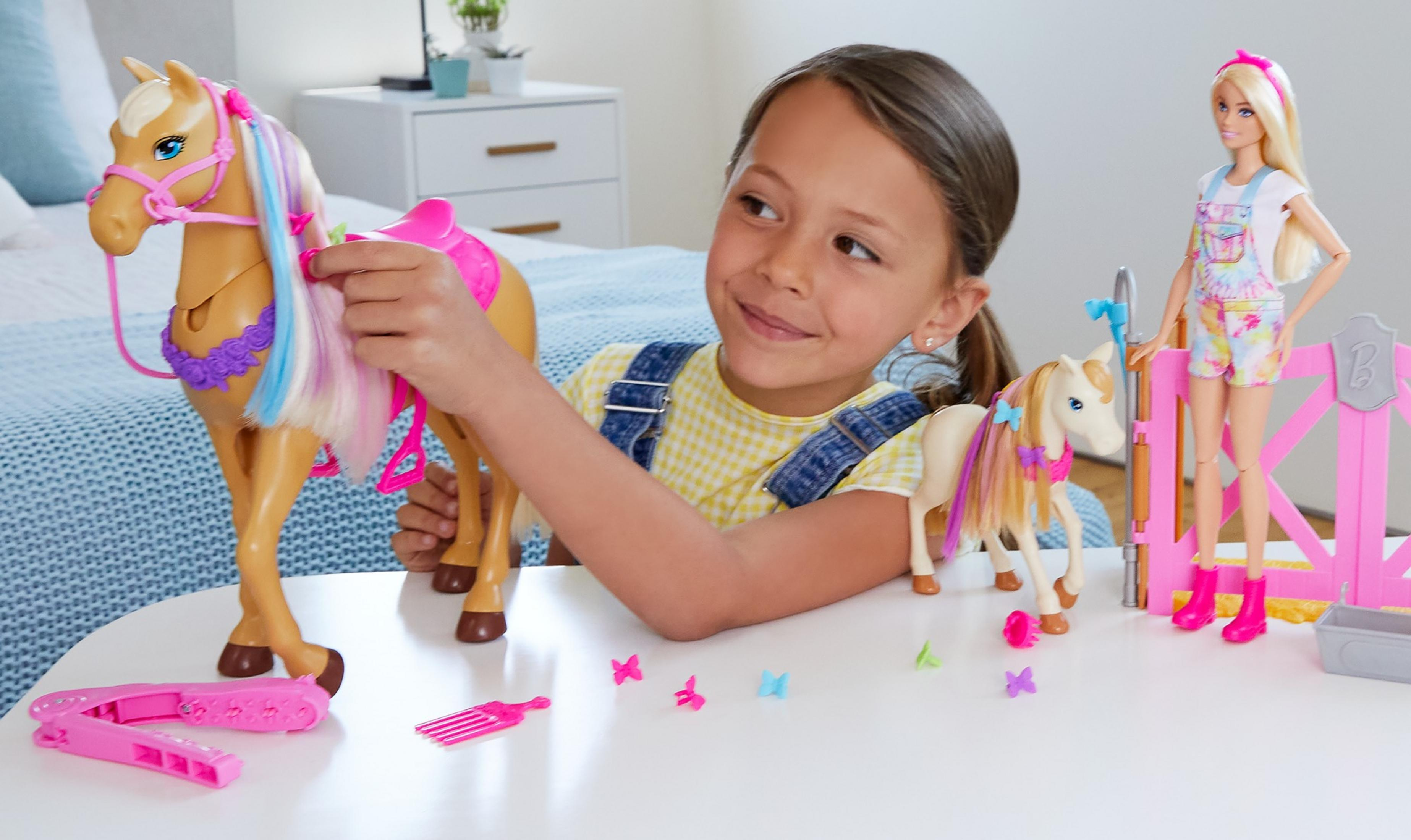 Dziewczynka bawi się zabawką koniem. Lalka Barbie i akcesoria na stole. W tle łóżko.