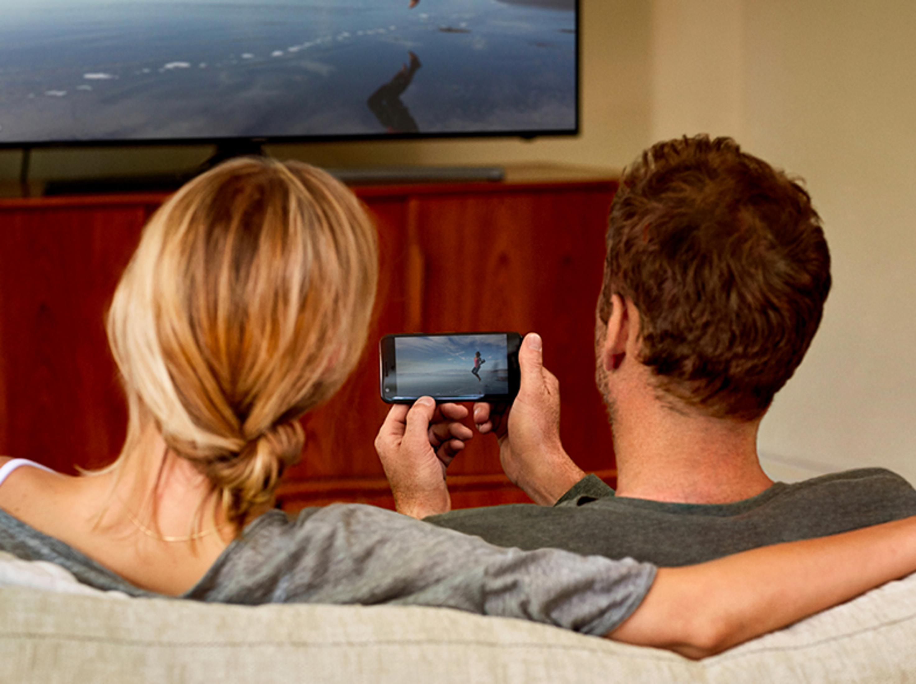 Ein Paar schaut ein Video auf einem Handy, mit einem Fernseher im Hintergrund.