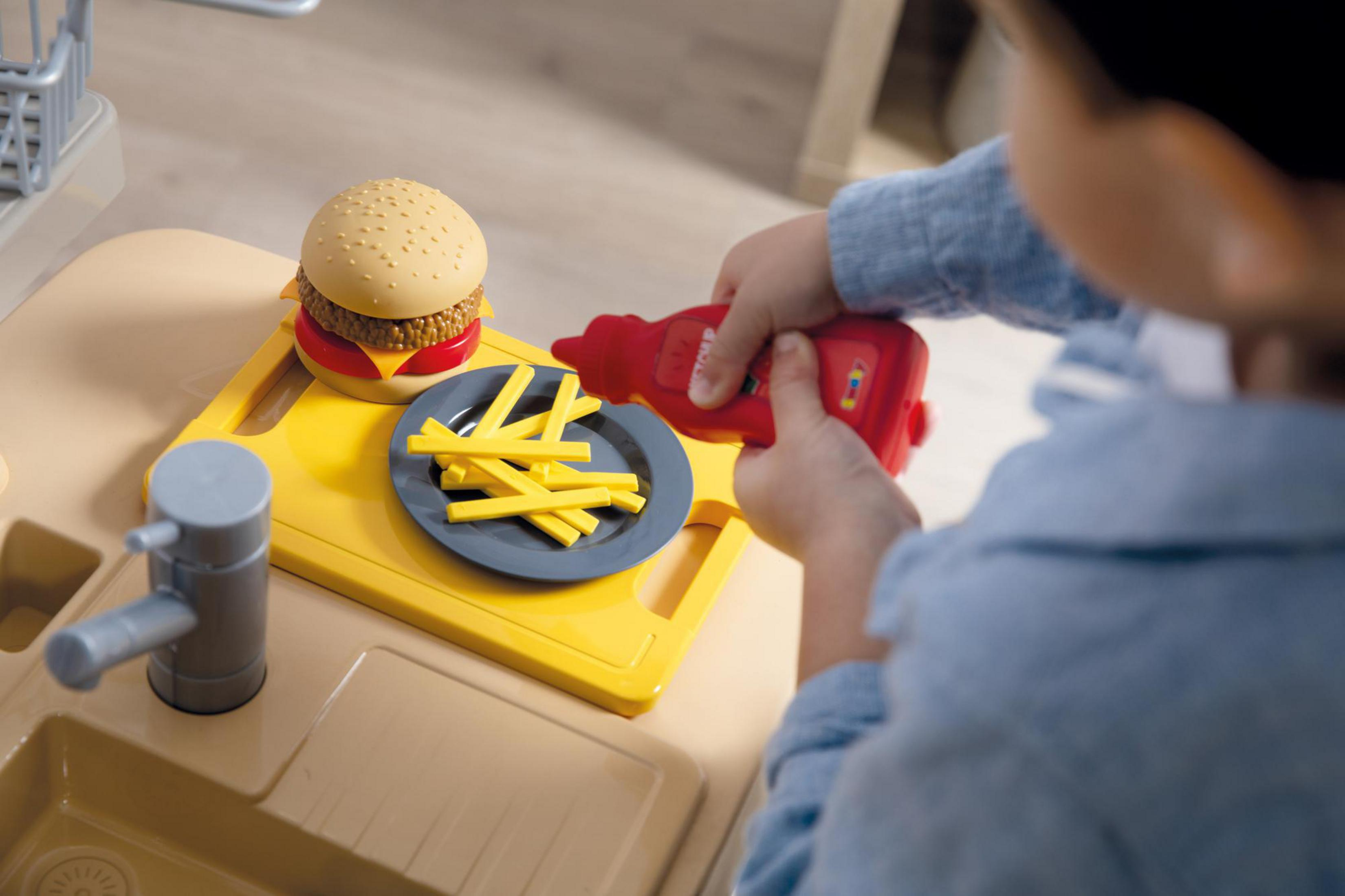 Dziecko wyciska ketchup na zabawkowych frytkach obok burgera na żółtej desce do krojenia.
