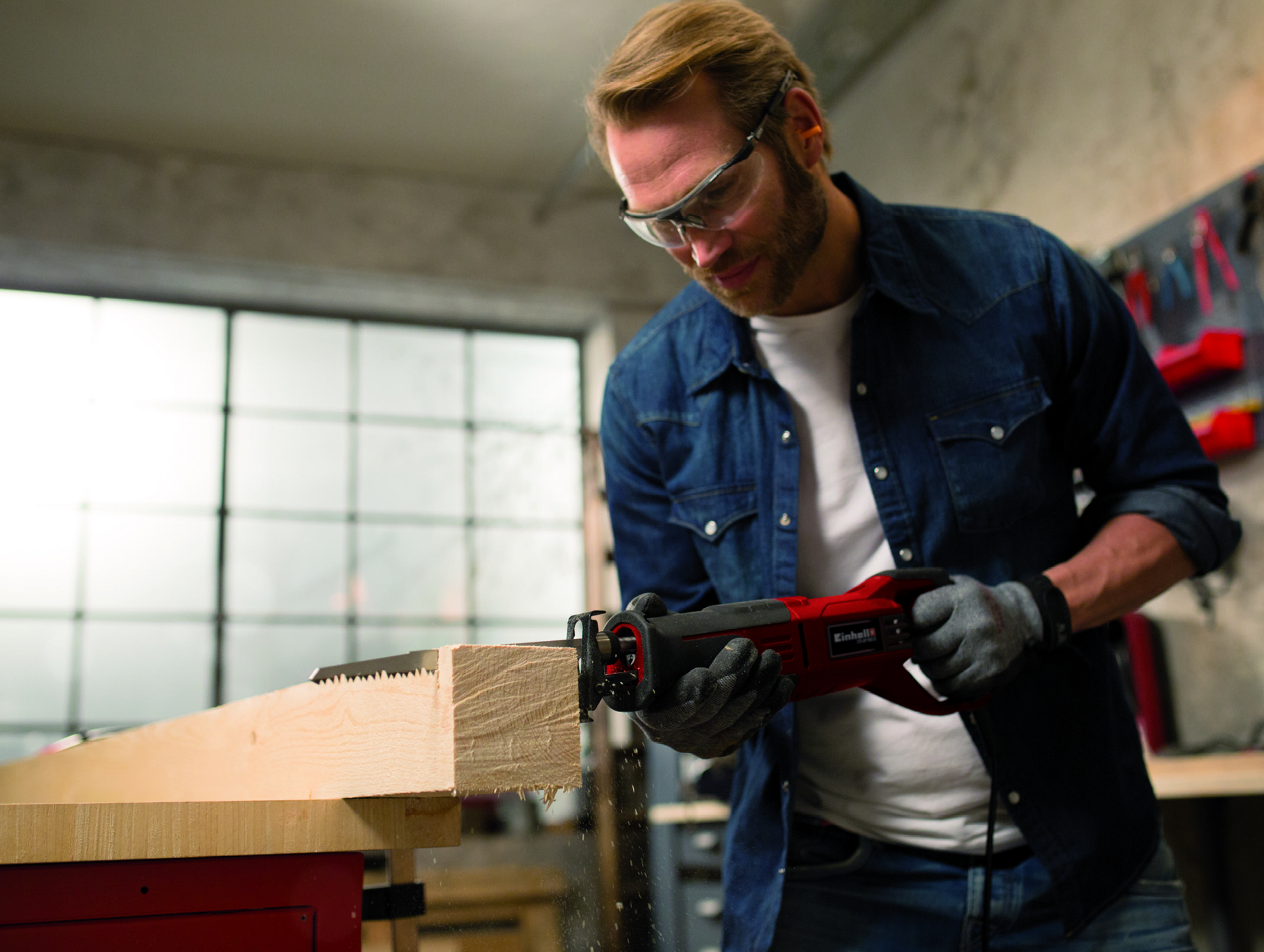 Hombre en un taller usando una sierra recíproca para cortar madera. Gafas de seguridad para protección.