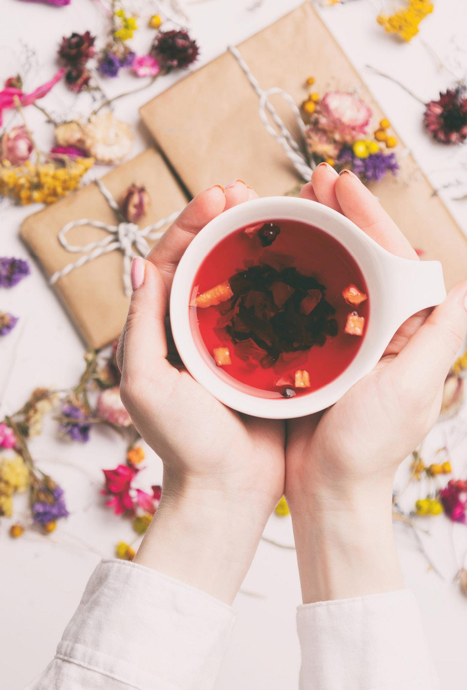 Hände halten eine weiße Tasse mit Tee. Zwei eingewickelte Geschenke und getrocknete Blumen im Hintergrund.