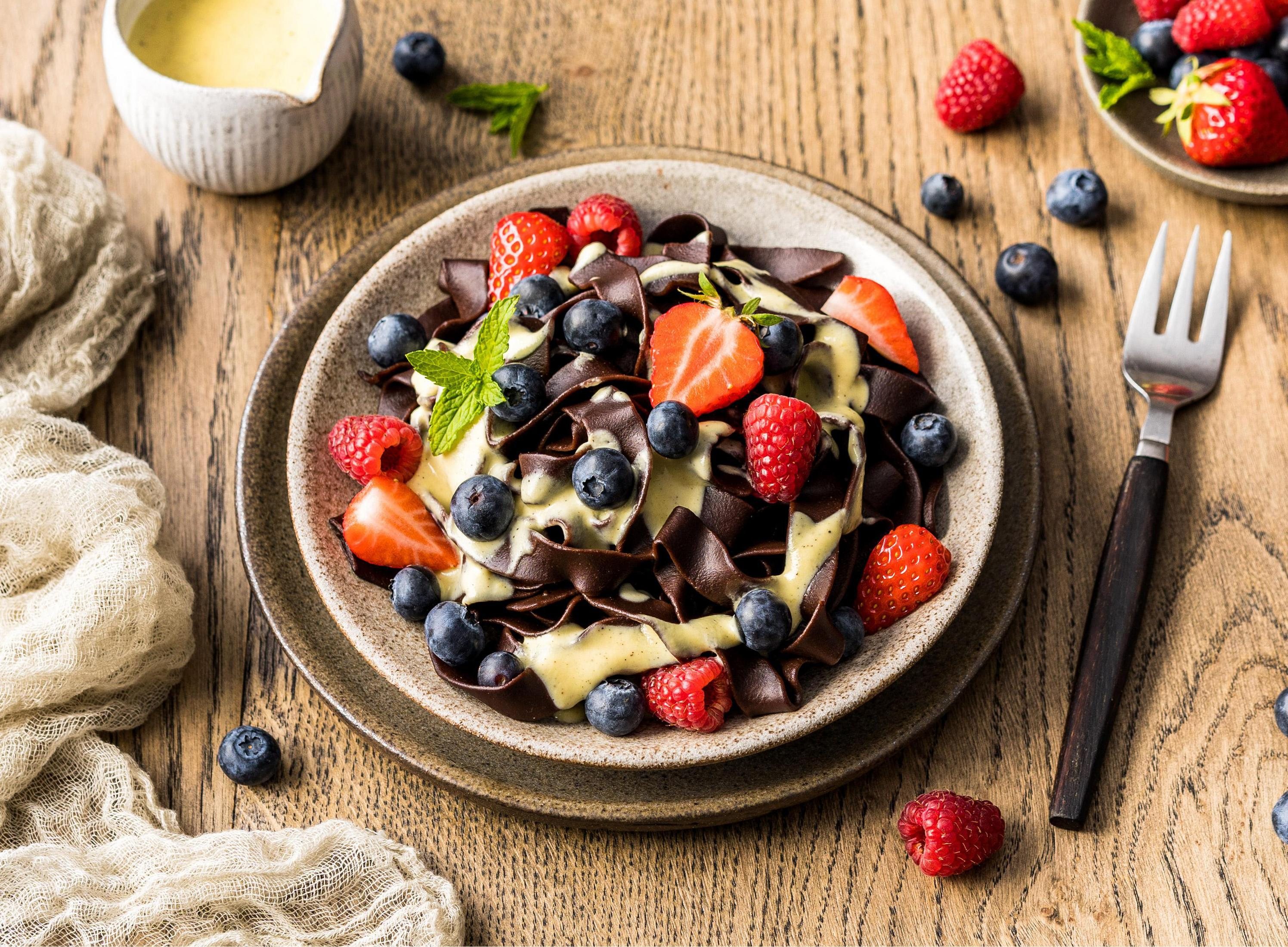 Danie z makaronem czekoladowym z jagodami i sosem. Biała miska i widelec na stole.