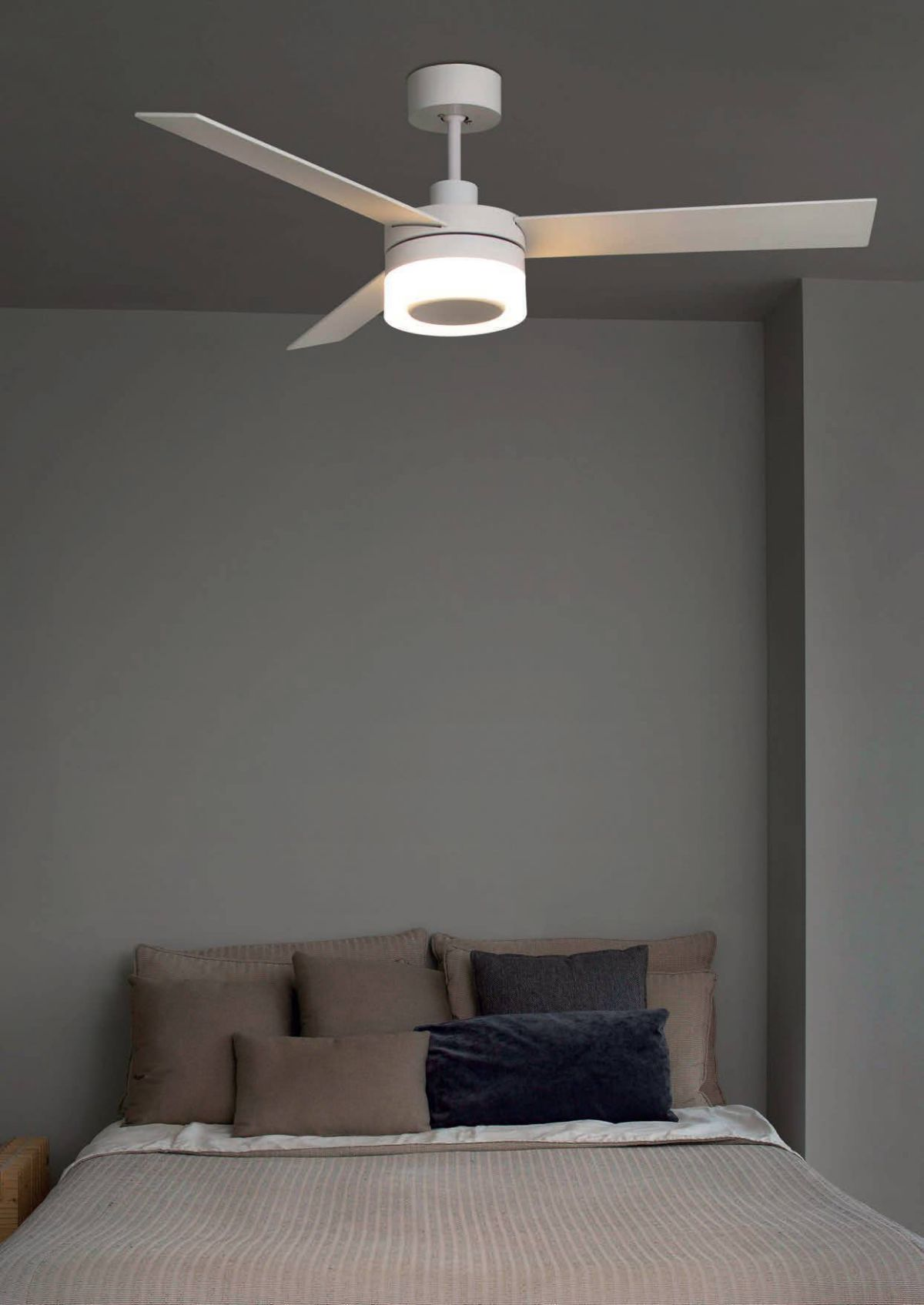 Ein weißer Deckenventilator mit drei Flügeln, der über einem Bett hängt. Die Wand ist grau.