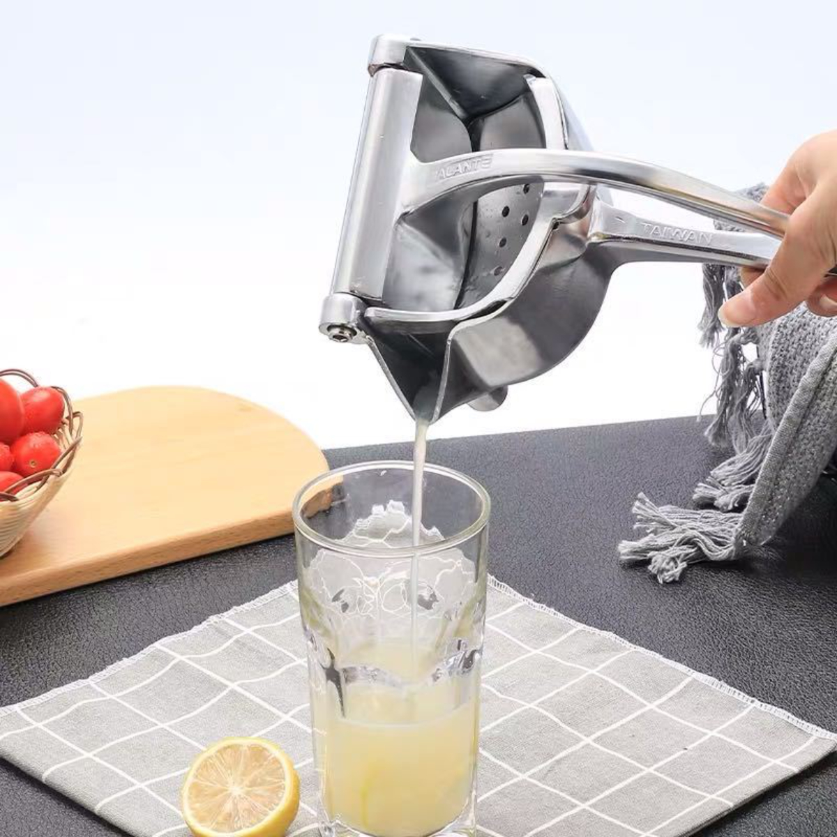 Silberne Saftpresse gießt Saft in ein Glas. Eine Zitrone und Tomaten sind auf dem Tisch.