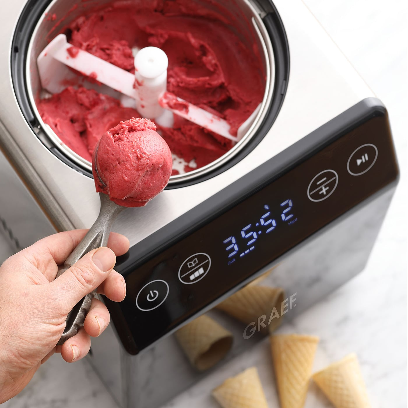 Eine Hand schöpft rosa Eis aus einer Maschine. Kegel sind im Vordergrund.