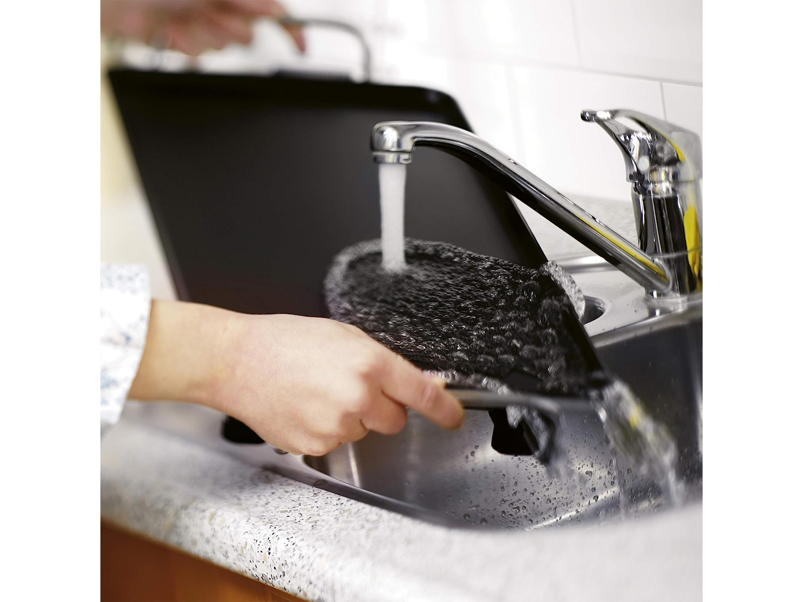 Person wäscht eine schwarze Grillpfanne in einem Waschbecken mit fließendem Wasser.