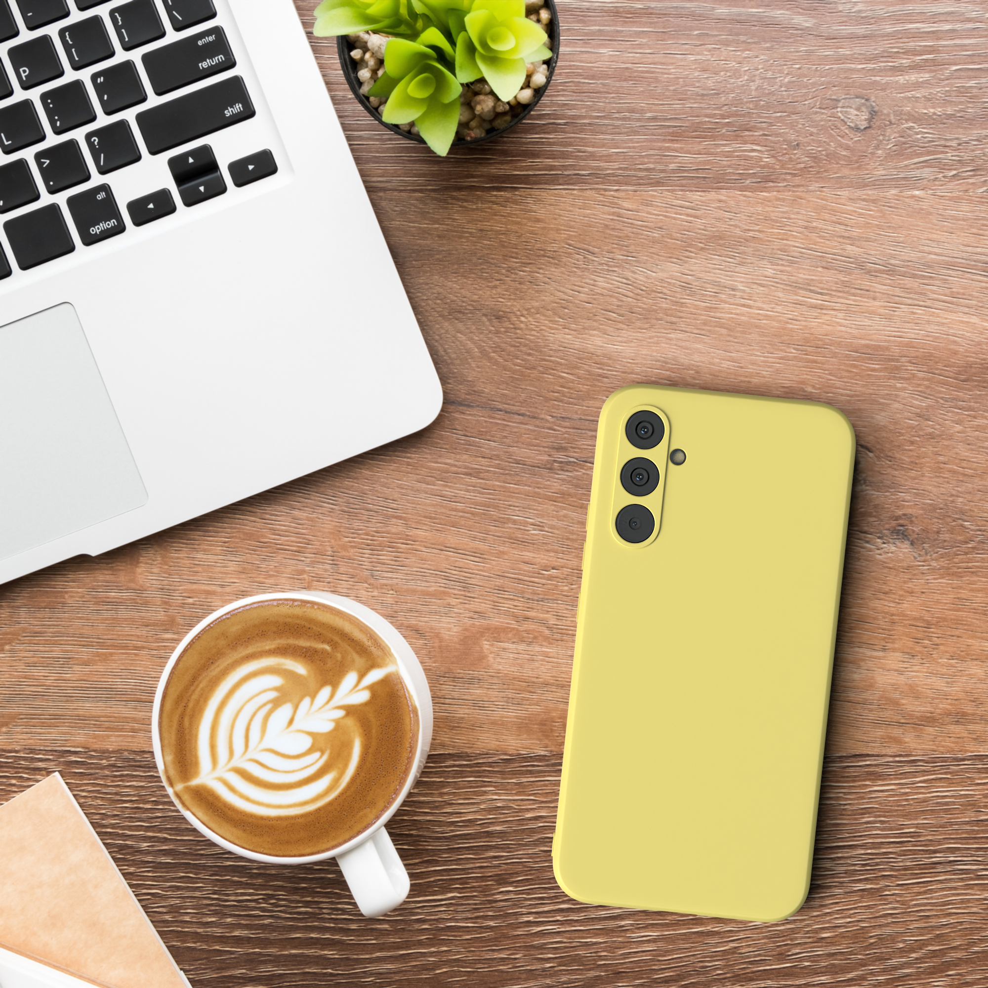 Ein Laptop, eine Tasse Kaffee, eine Handyhülle und eine kleine Pflanze auf einem Holztisch. Die Handyhülle ist gelb.