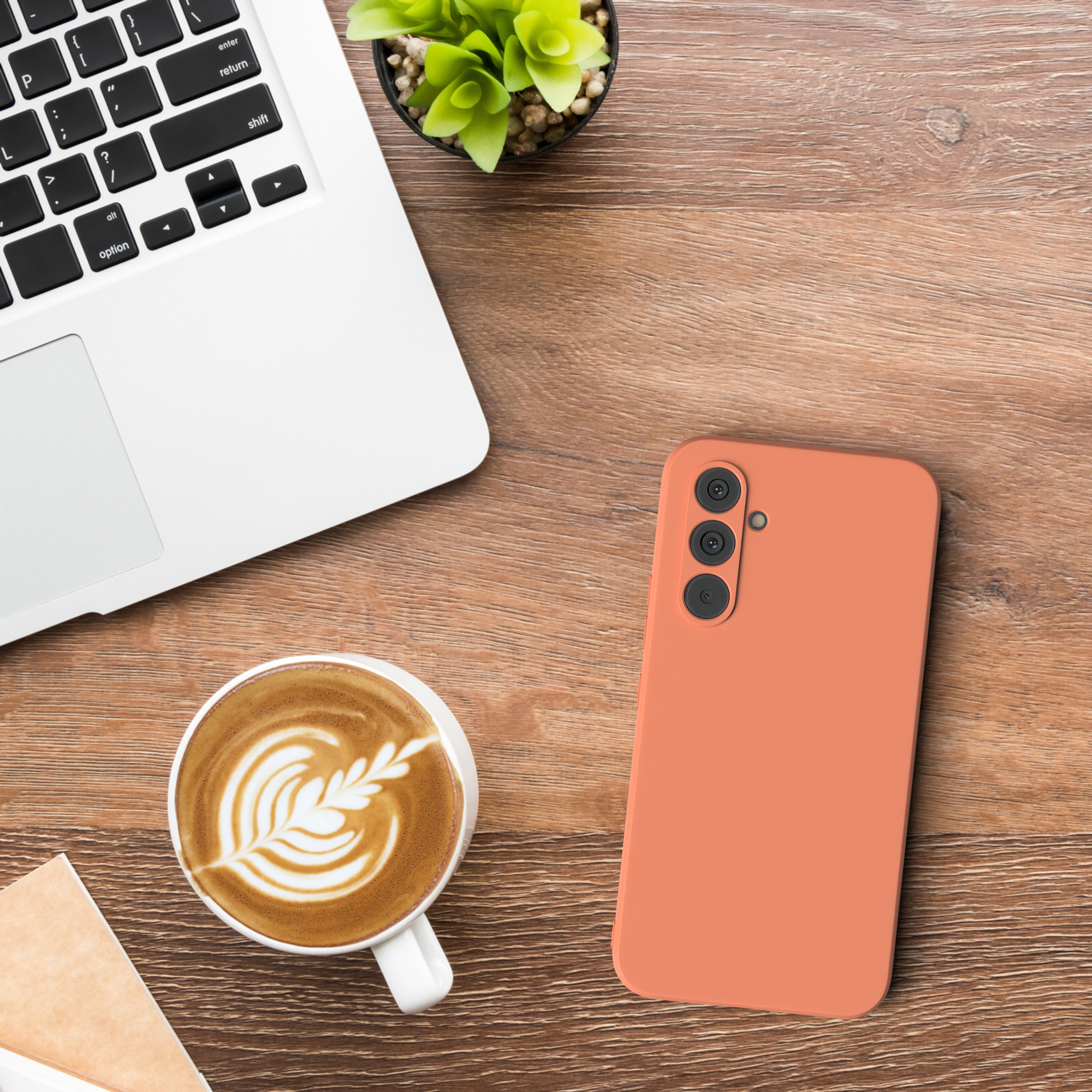 Draufsicht: Laptop, Kaffee mit Latte Art, orangefarbene Handyhülle und eine kleine Pflanze auf einem Holztisch.