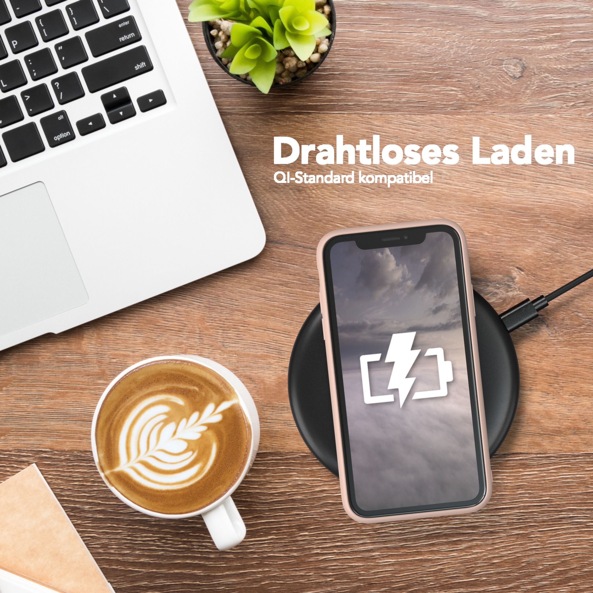 Telefon wird auf einem Pad aufgeladen. Kaffeetasse und Laptop auf einem Holztisch, Pflanze im Hintergrund.