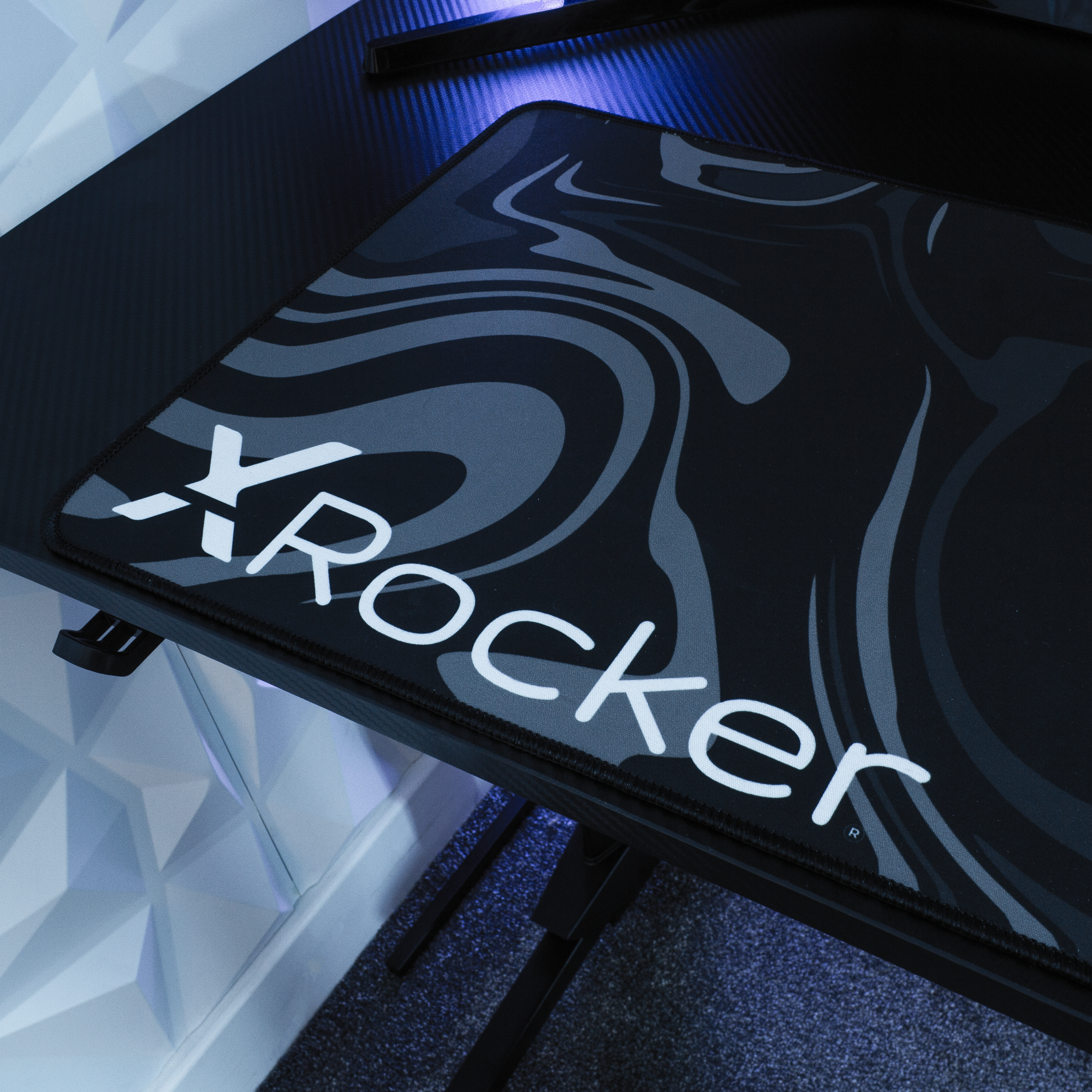 Ein schwarzer Schreibtisch mit einer Mausunterlage mit dem weißen Wort „X Rocker“ darauf. Der Schreibtisch ist dunkel.