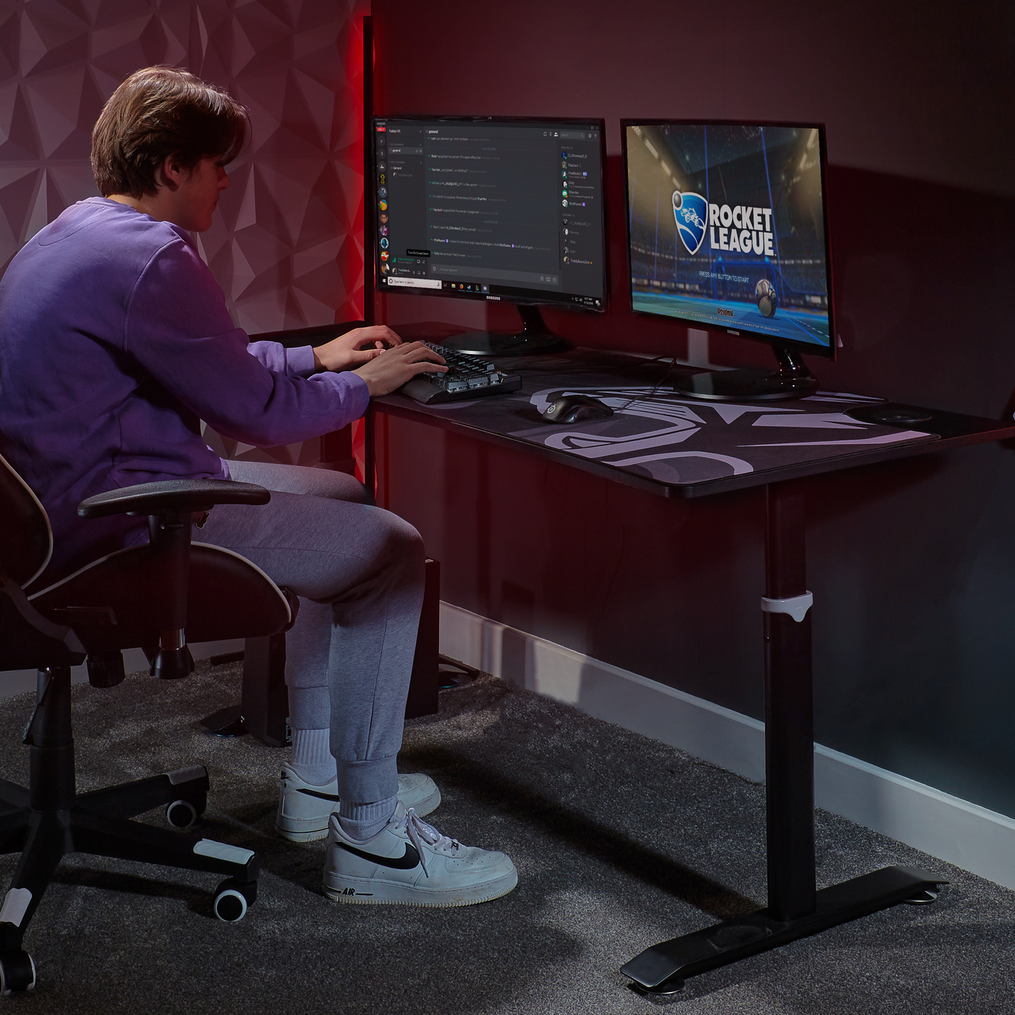 Eine Person spielt an einem Schreibtisch mit zwei Monitoren. Tastatur und Maus sind sichtbar. Der Raum ist dunkel.
