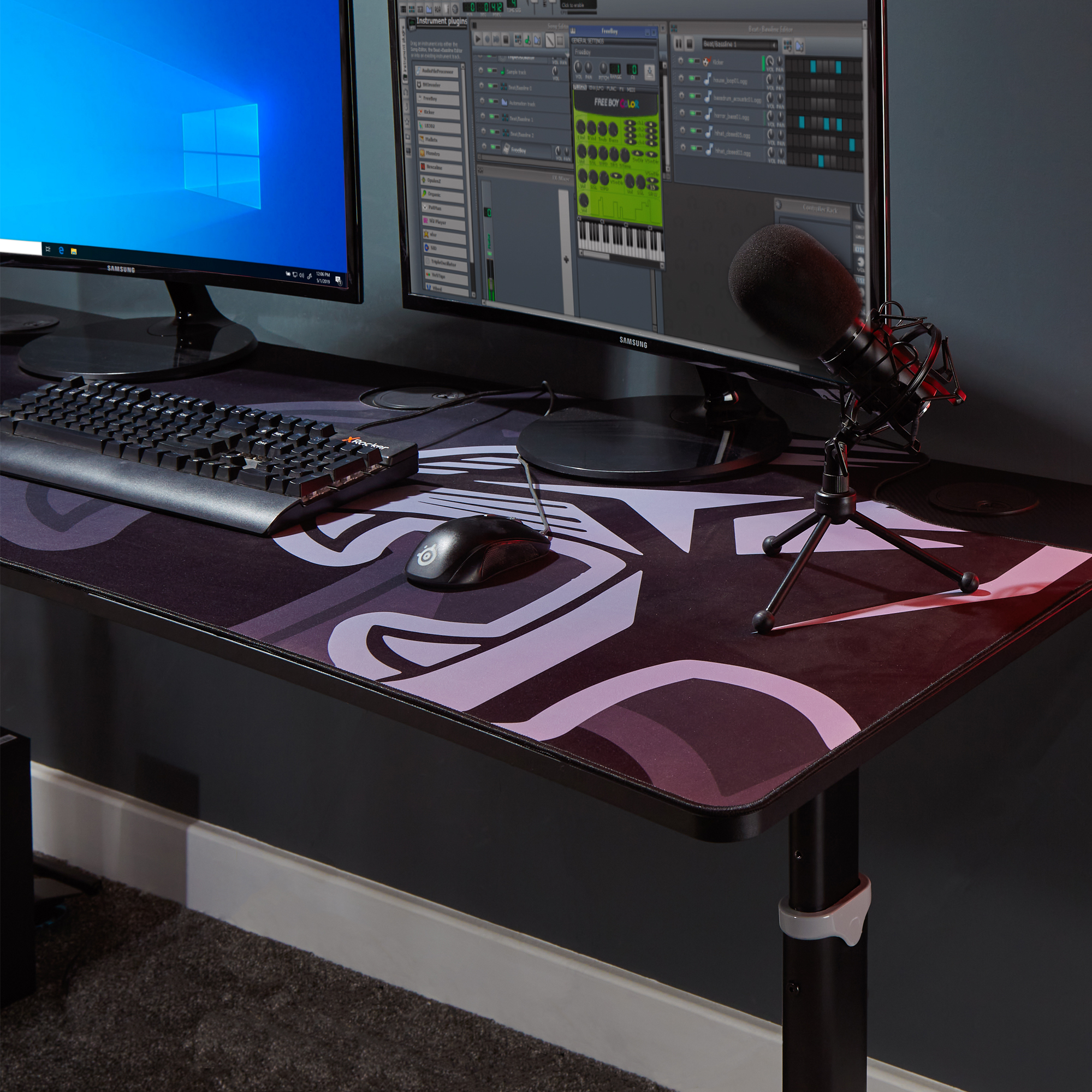 Ein schwarzer Schreibtisch mit Tastatur, Maus und Mikrofon. Zwei Monitore im Hintergrund.