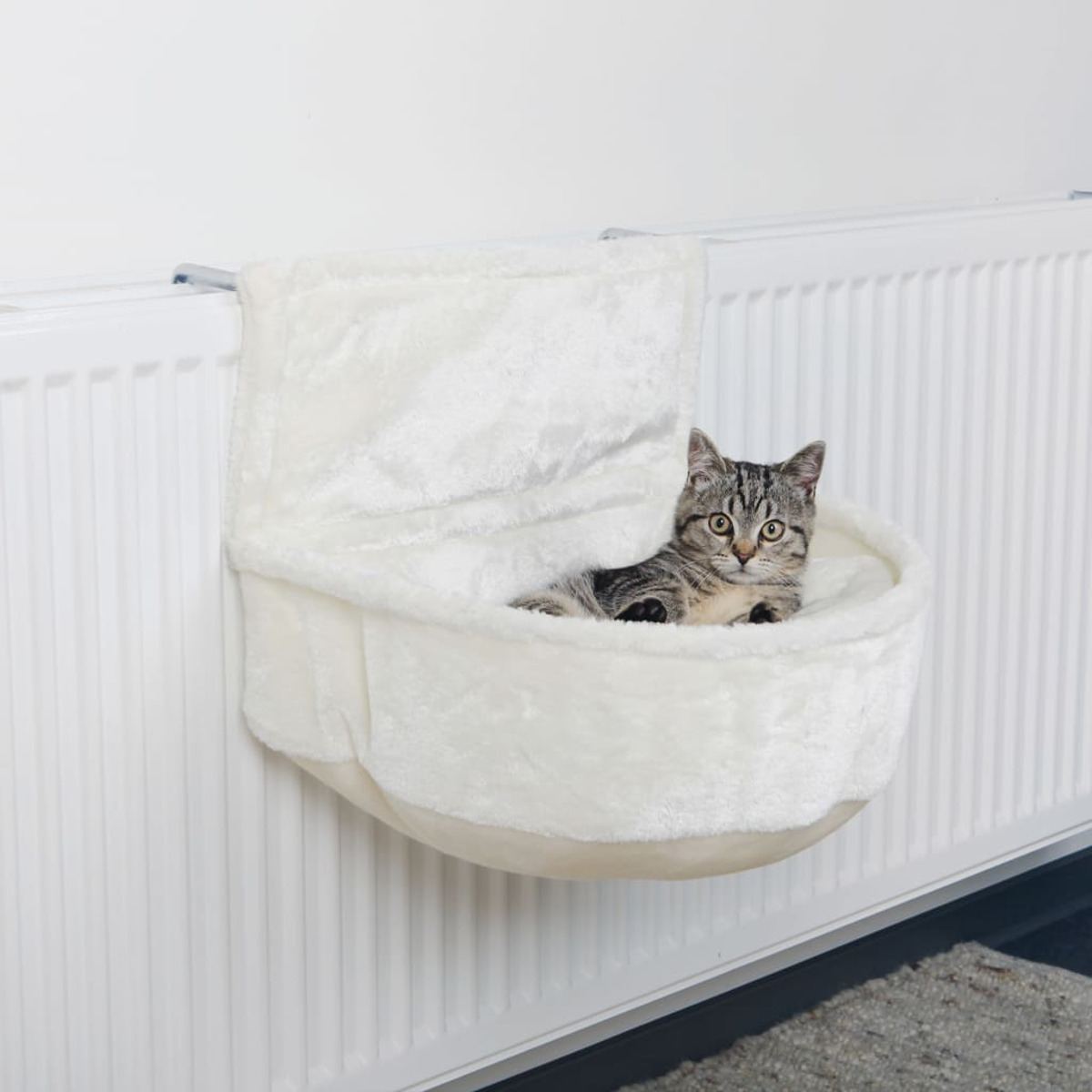 Eine Katze ruht in einem Katzenbett auf einem Heizkörper. Das Bett ist weiß und flauschig.