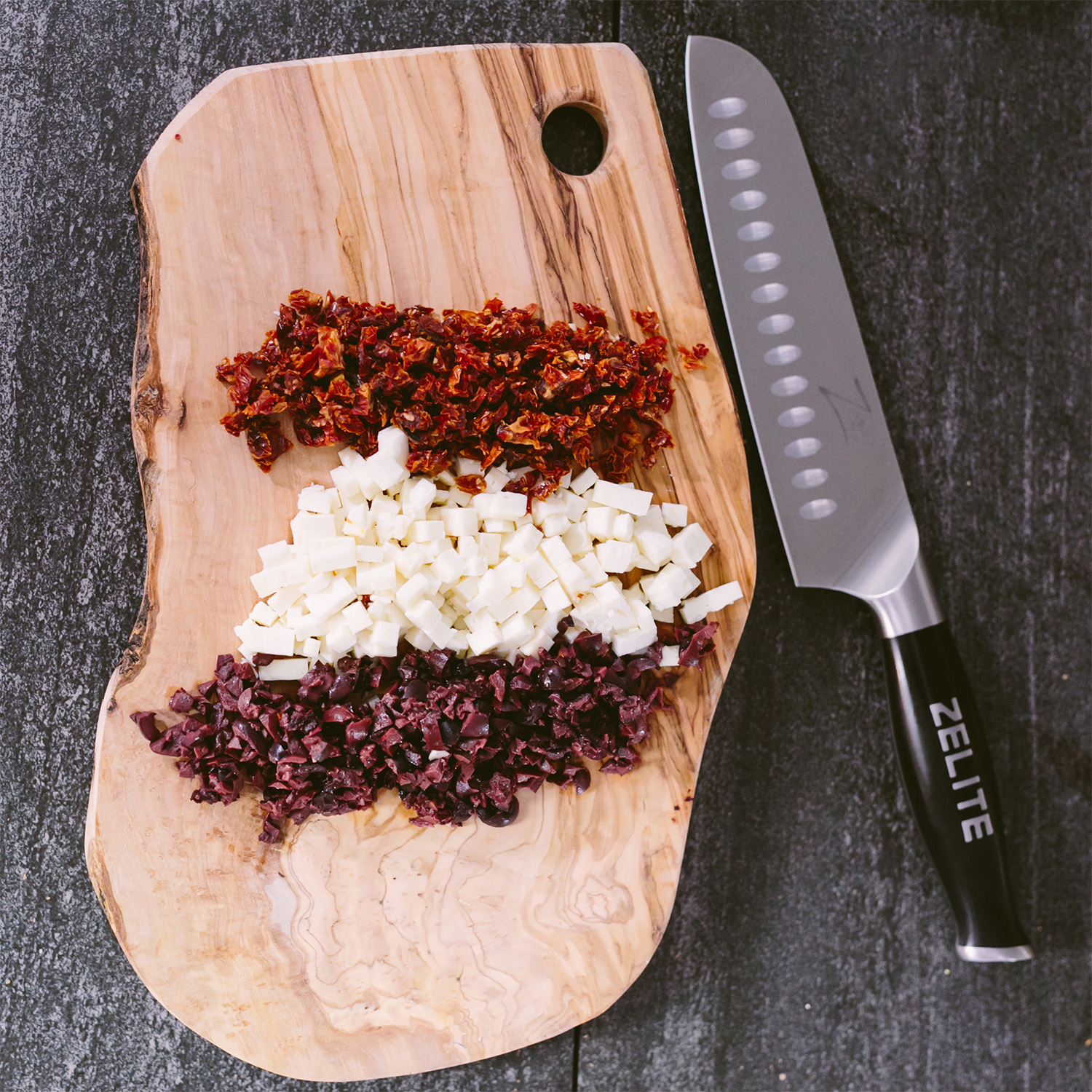 Holzschneidebrett mit gehackten Tomaten, Käse und Oliven, daneben ein Messer.