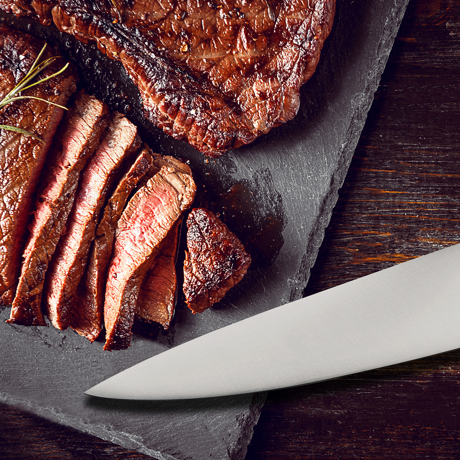 Geschnittenes Steak auf Schieferplatte, mit Messer und gegrilltem Steak im Hintergrund.