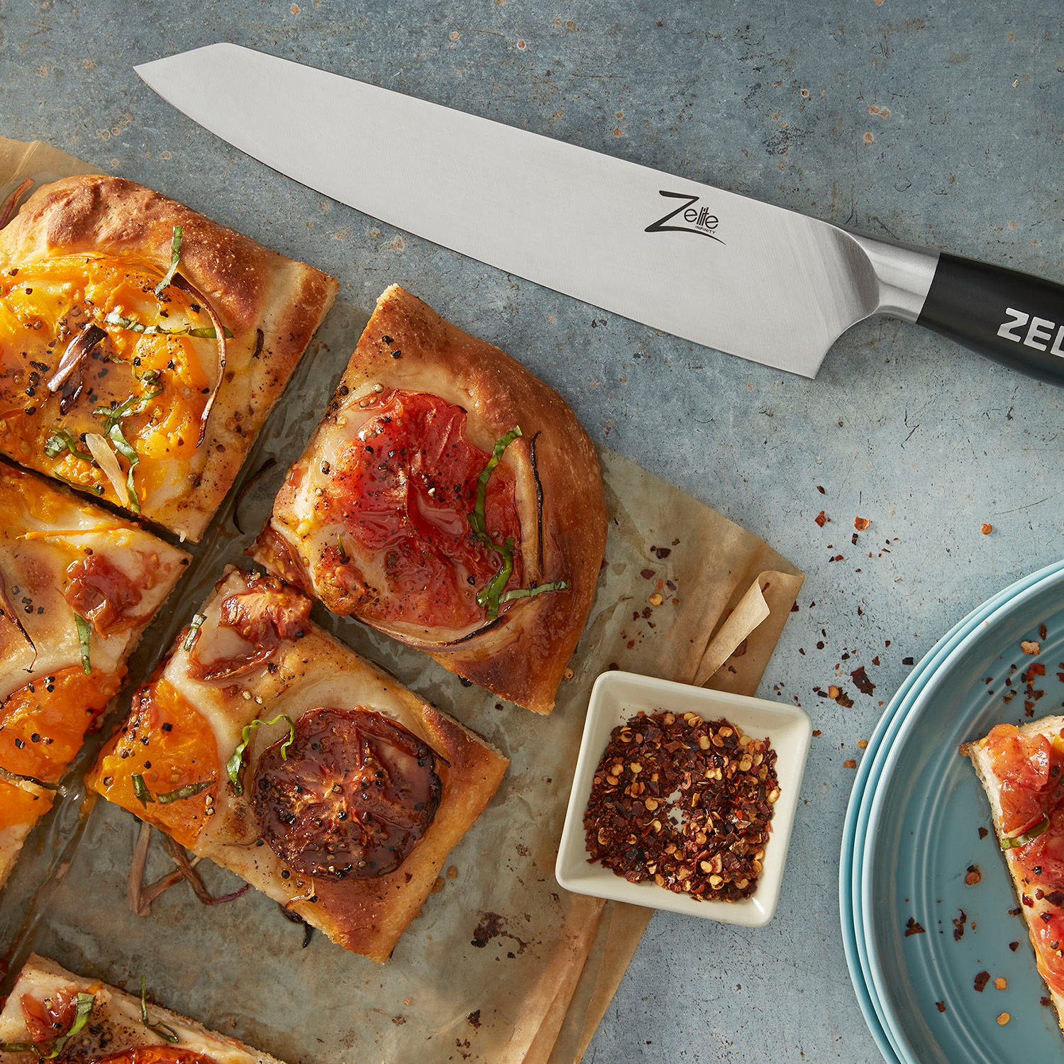 Ein Kochmesser neben geschnittener Pizza mit Gemüse und einer kleinen Schüssel mit roten Gewürzen.