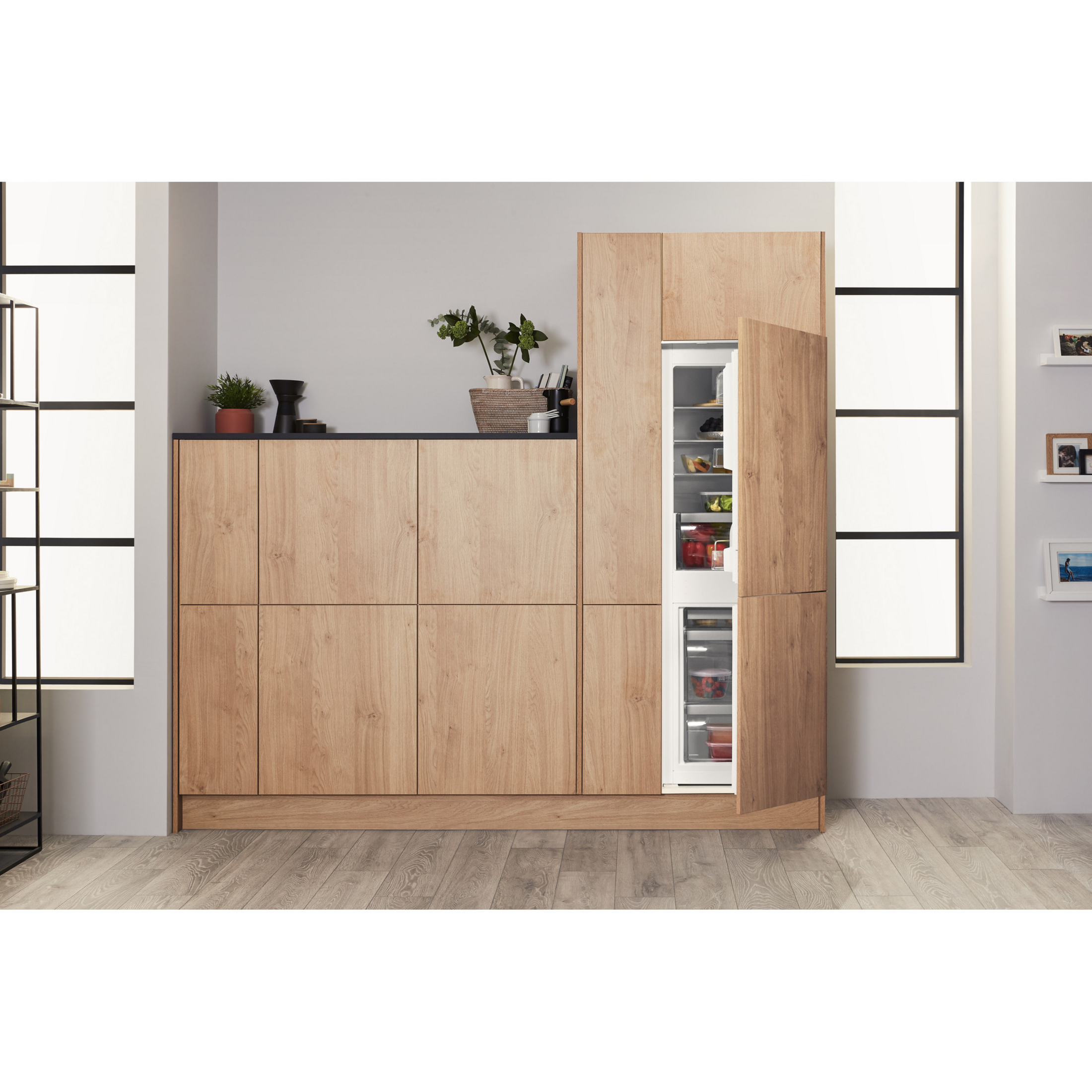 Küchenschränke, eine Tür offen, die einen Kühlschrank zeigt. Helle Holzschränke, weißes Innere und eine Pflanze.
