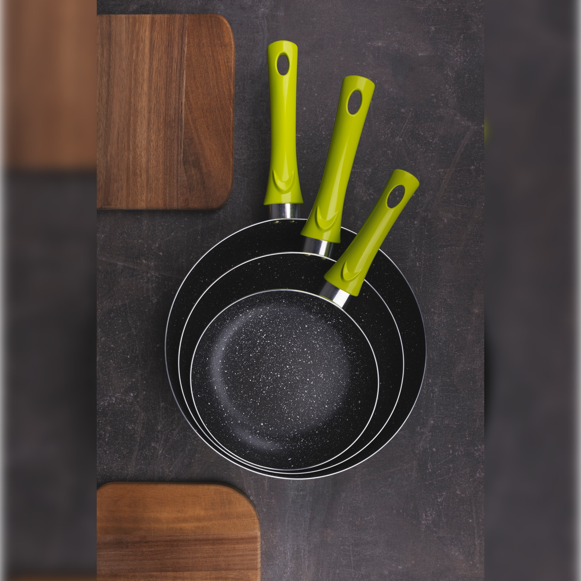 Tres sartenes negras anidadas con asas verdes y tablas de cortar de madera alrededor.