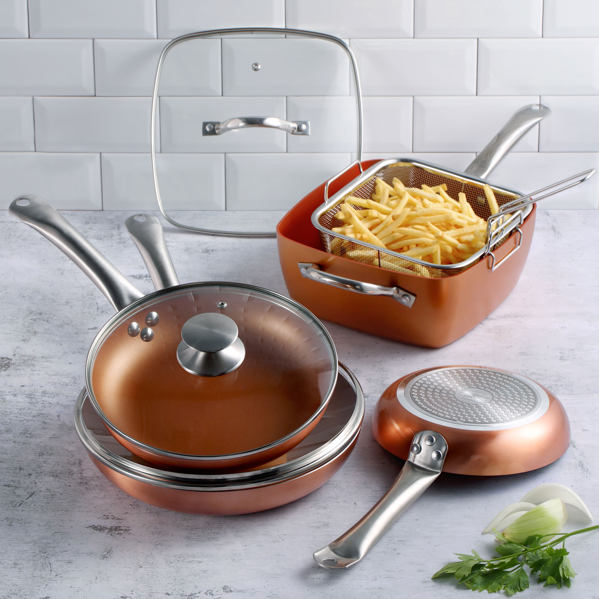 Kupferfarbene Töpfe und Pfannen mit Deckel, Frittierkorb mit Pommes und Pfannenwender auf weißer Oberfläche.