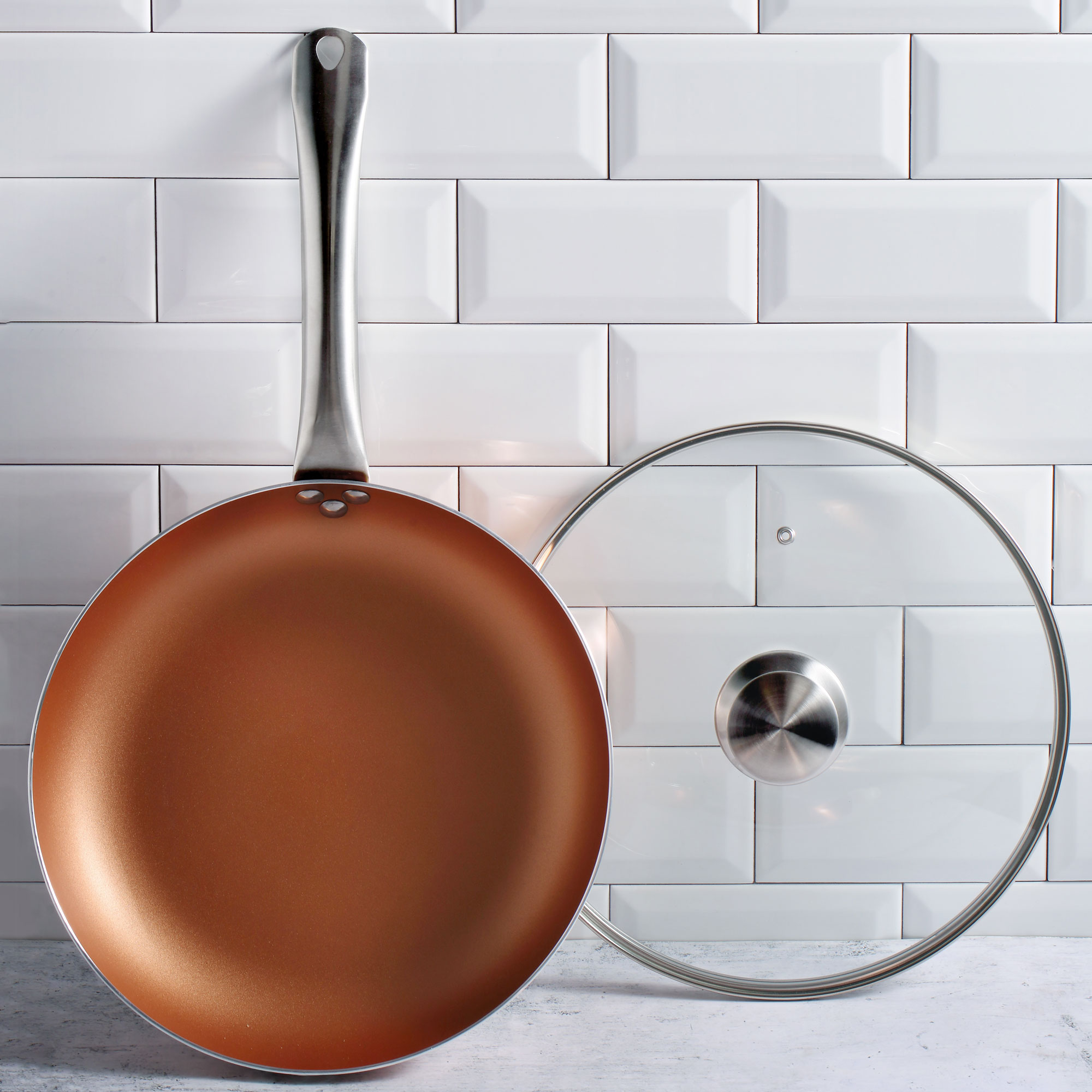 Sartén de color cobre con tapa transparente. Ambos están sobre una superficie de baldosas blancas.