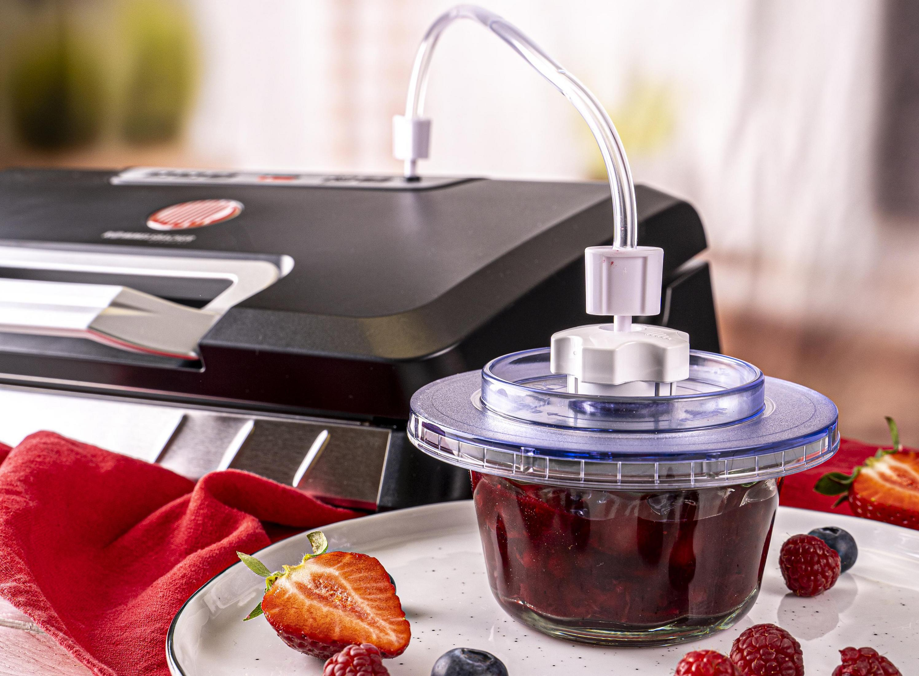 Maszyna do pakowania próżniowego ze słoikiem czerwonego dżemu i jagodami na talerzu. Czerwona serwetka.