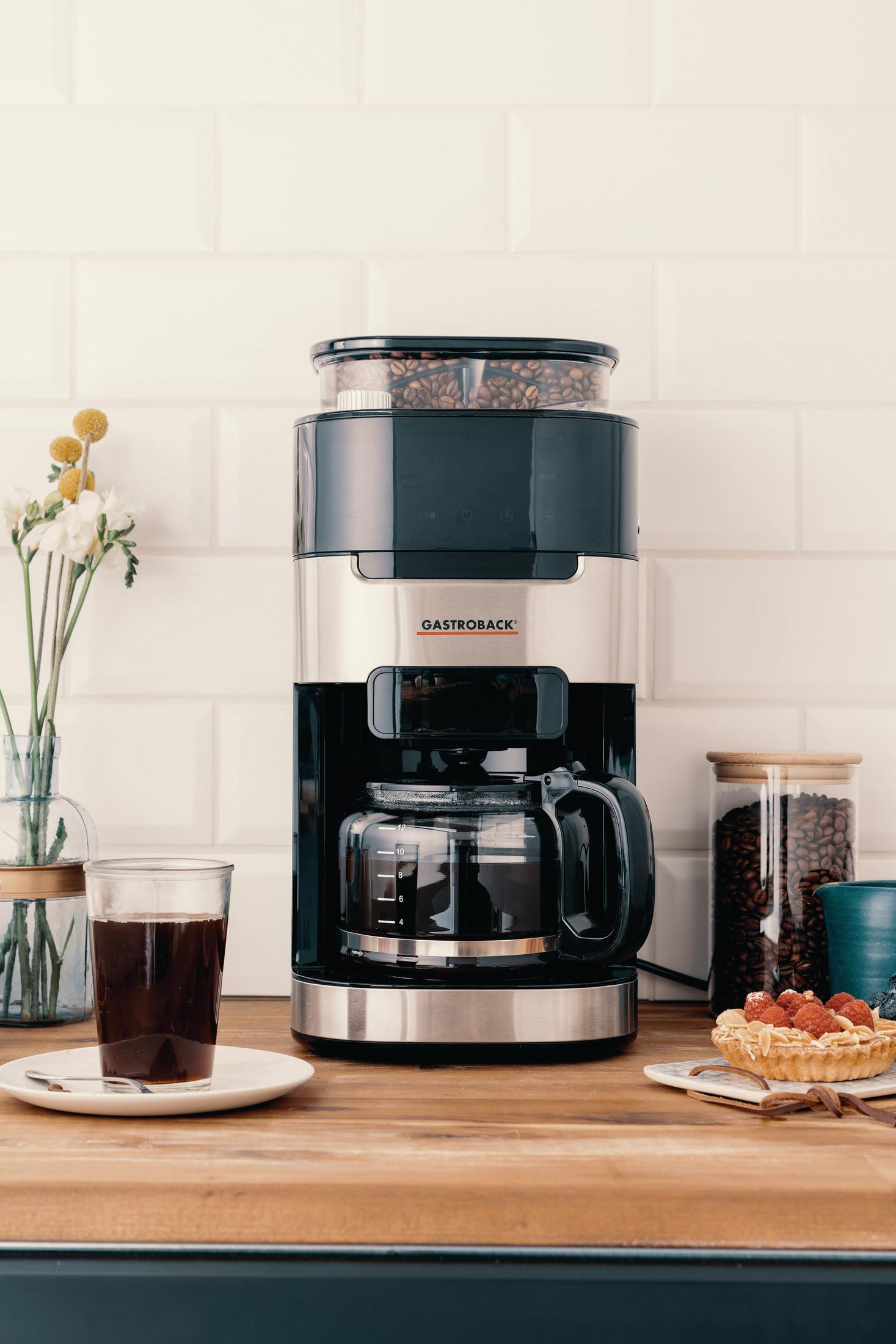 Ekspres do kawy na drewnianym blacie. Kawa, kwiaty i tarty na blacie. Białe tło z płytek.