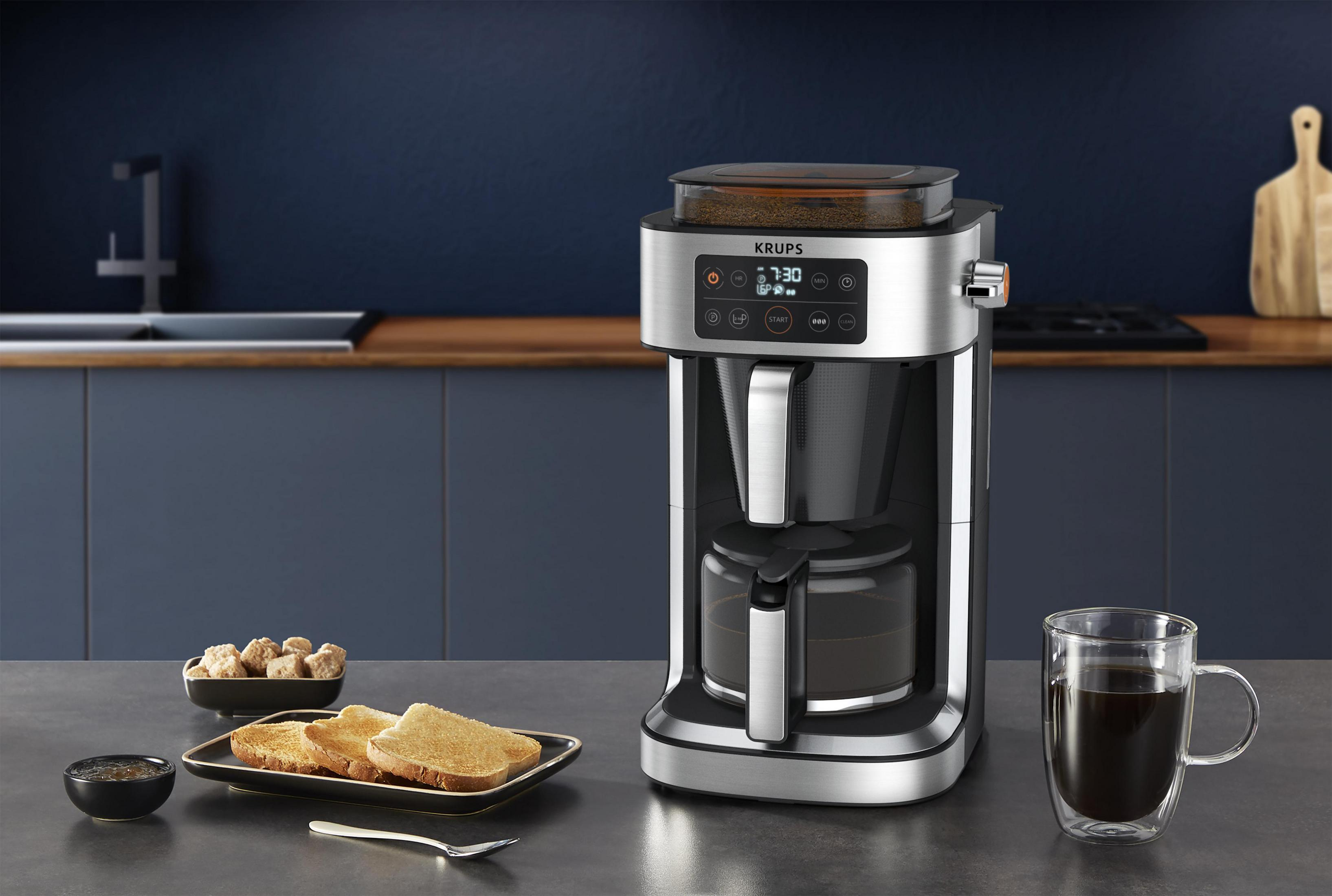 Ekspres do kawy ze szklaną karafką, tosty, kostki cukru i filiżanka kawy na szarym blacie.