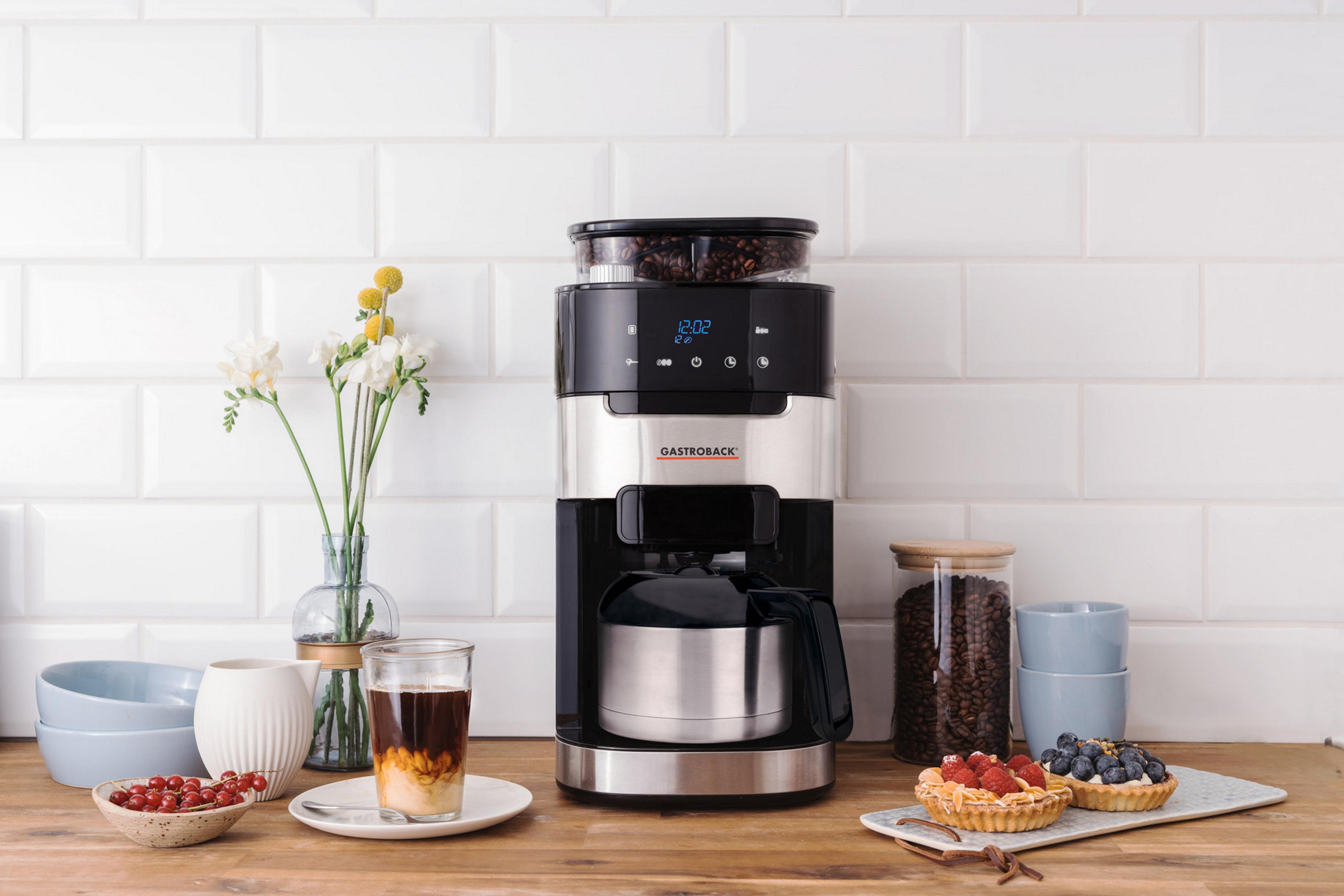 Macchina da caffè su bancone in legno con caffè, crostate, fiori e sfondo piastrellato bianco.