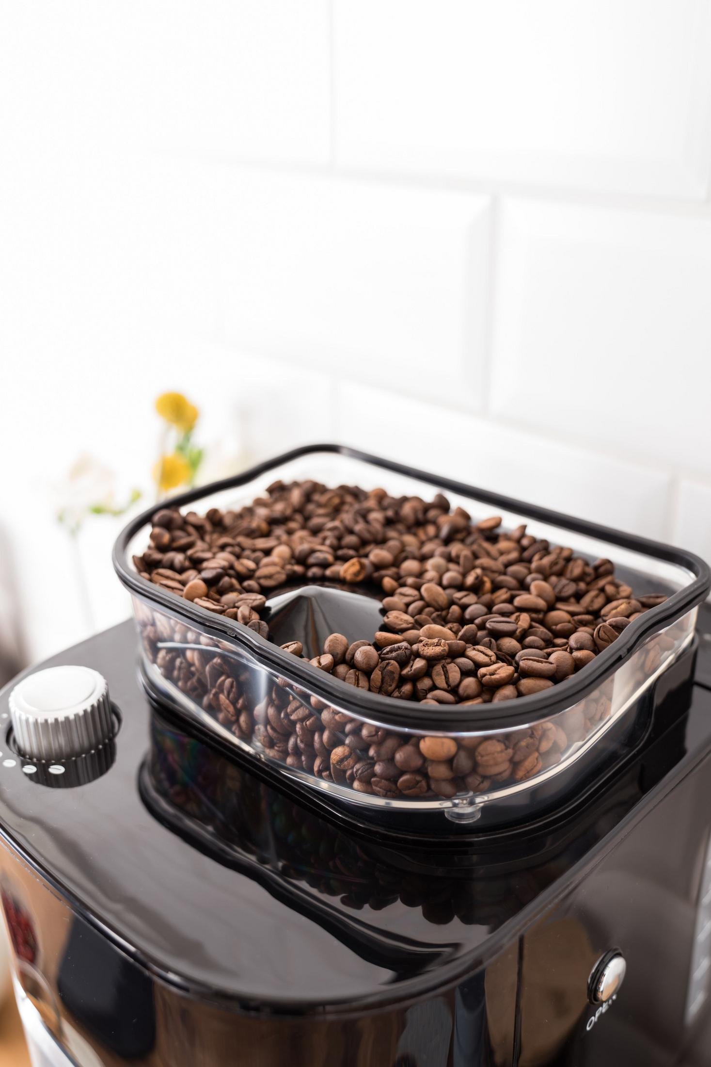 Chicchi di caffè in un contenitore trasparente su una macchina da caffè nera. Sfondo di piastrelle bianche.