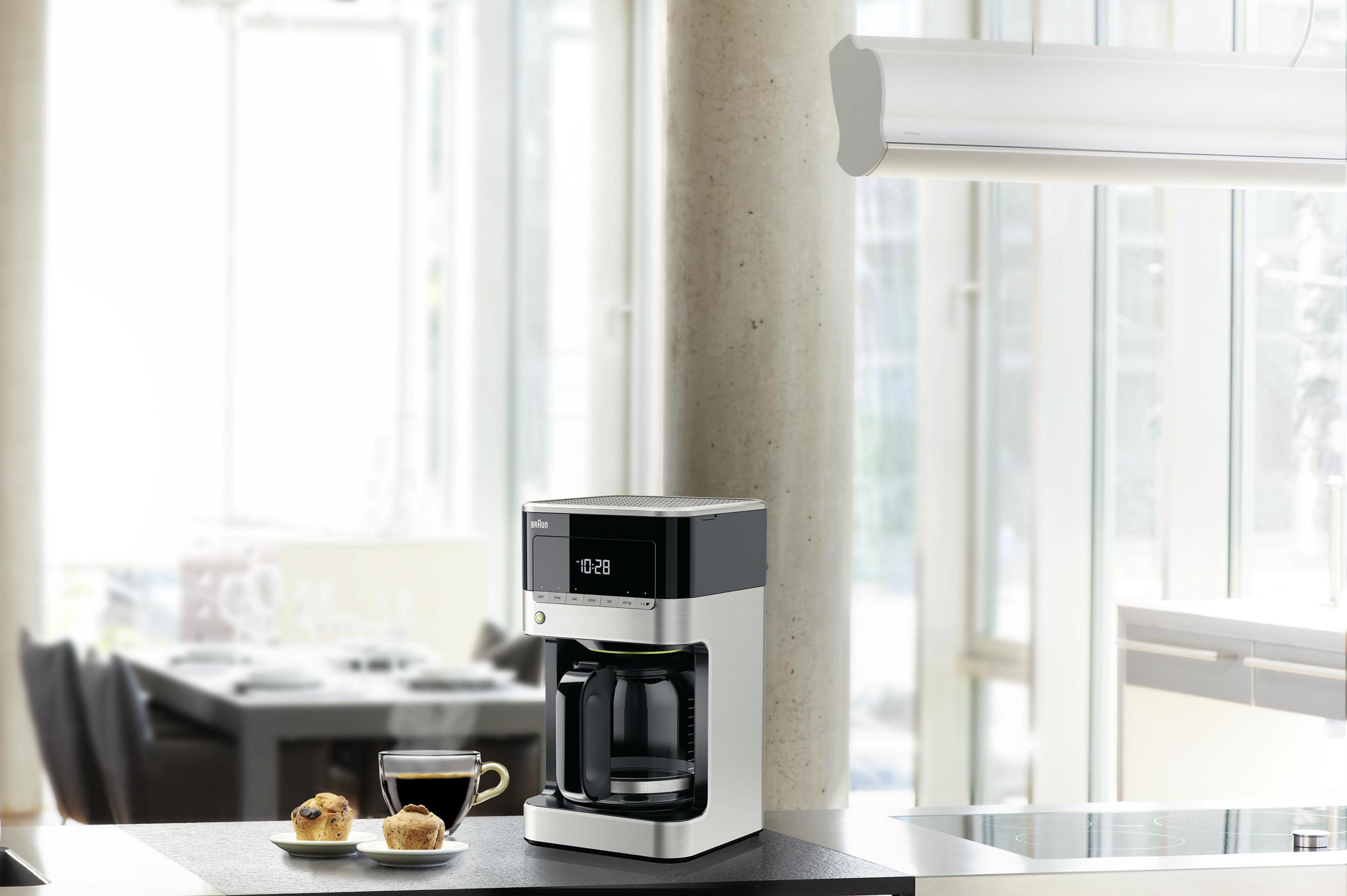 Ekspres do kawy na blacie kuchennym. W tle filiżanka kawy, dwa ciastka i okno.