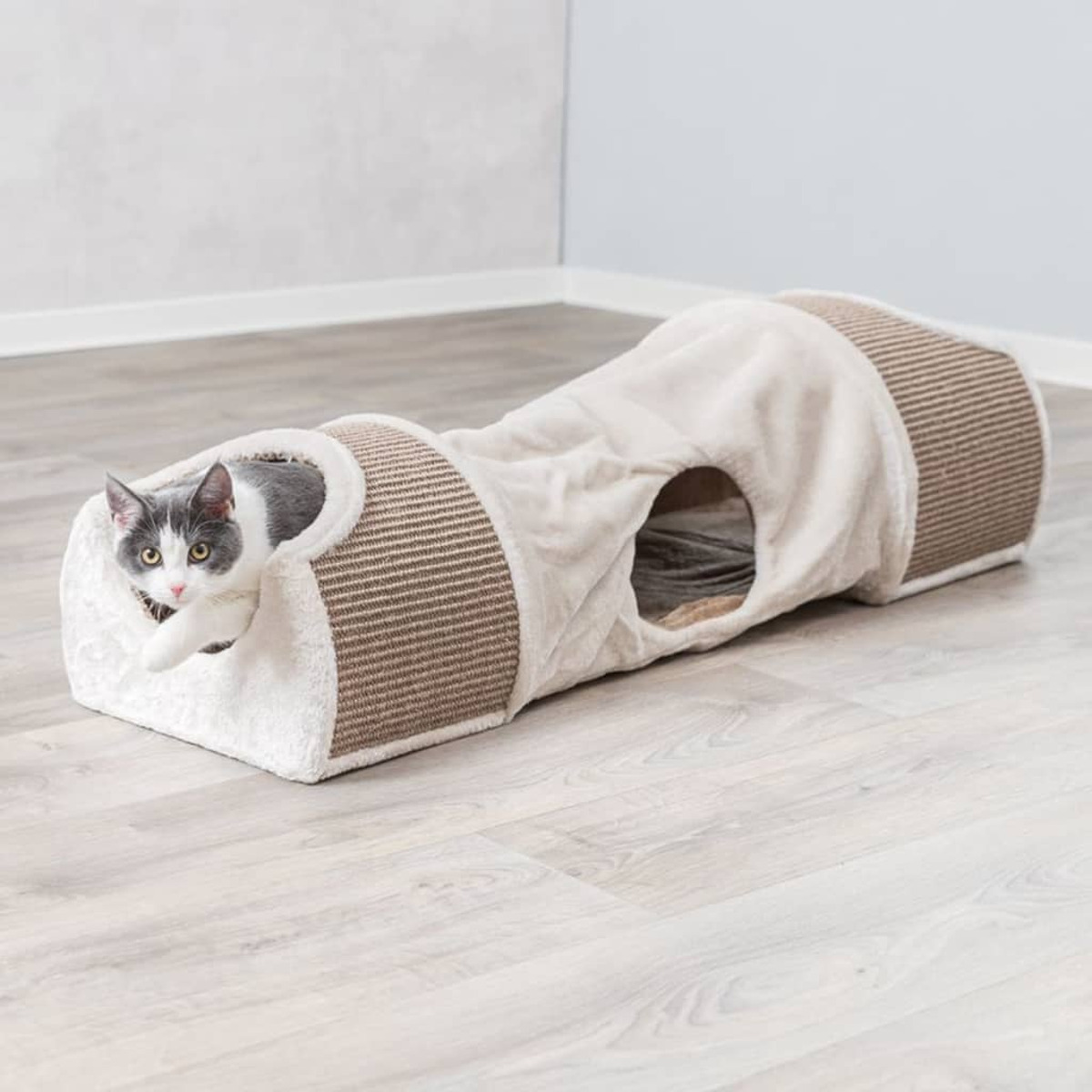 Eine Katze, die aus einem Katzentunnel aus beigem und braunem Material auf einem Holzboden schaut.